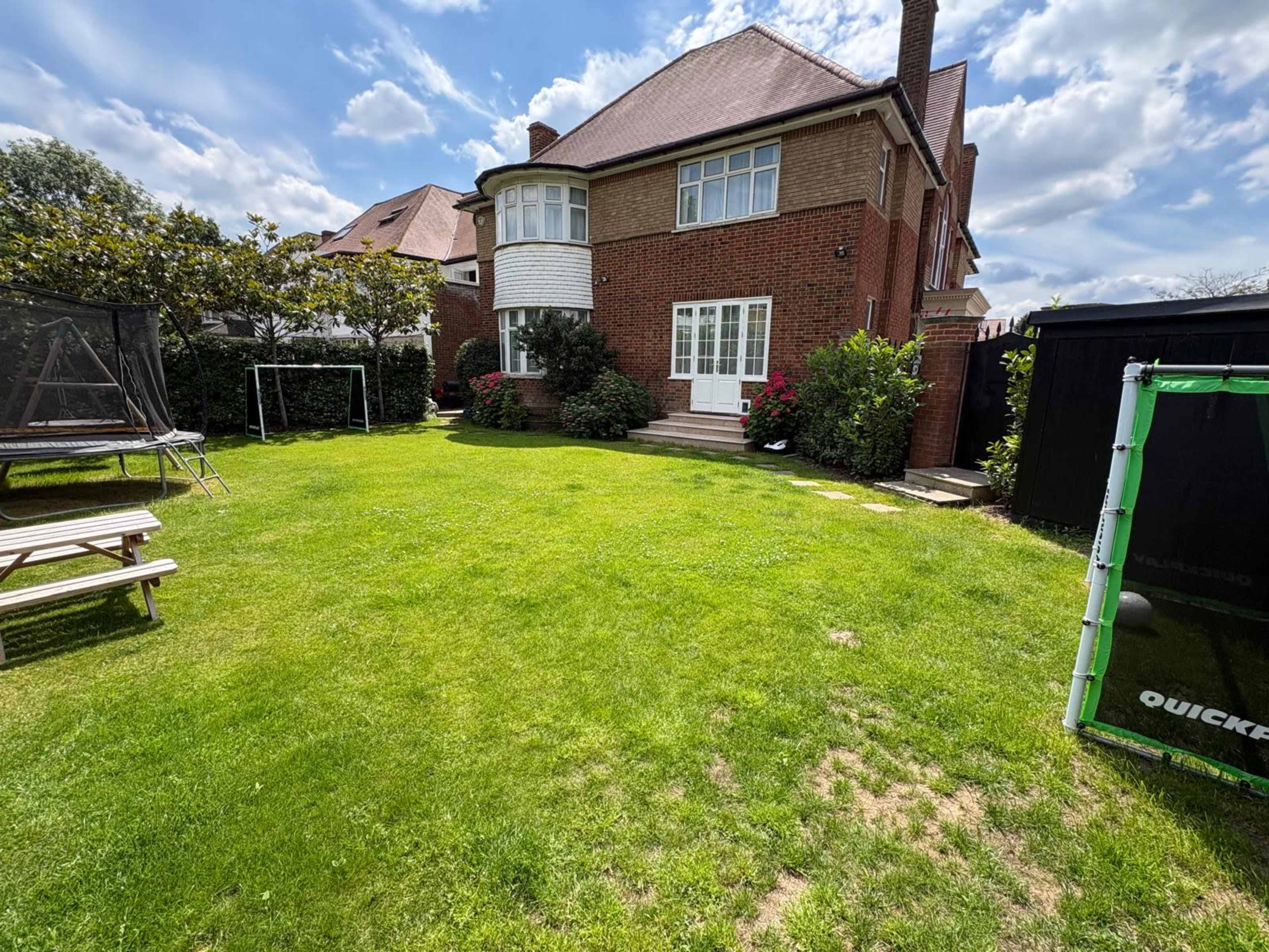 The image shows a well-maintained garden with a brick house, a trampoline, and a neat lawn surrounded by flowerbeds.