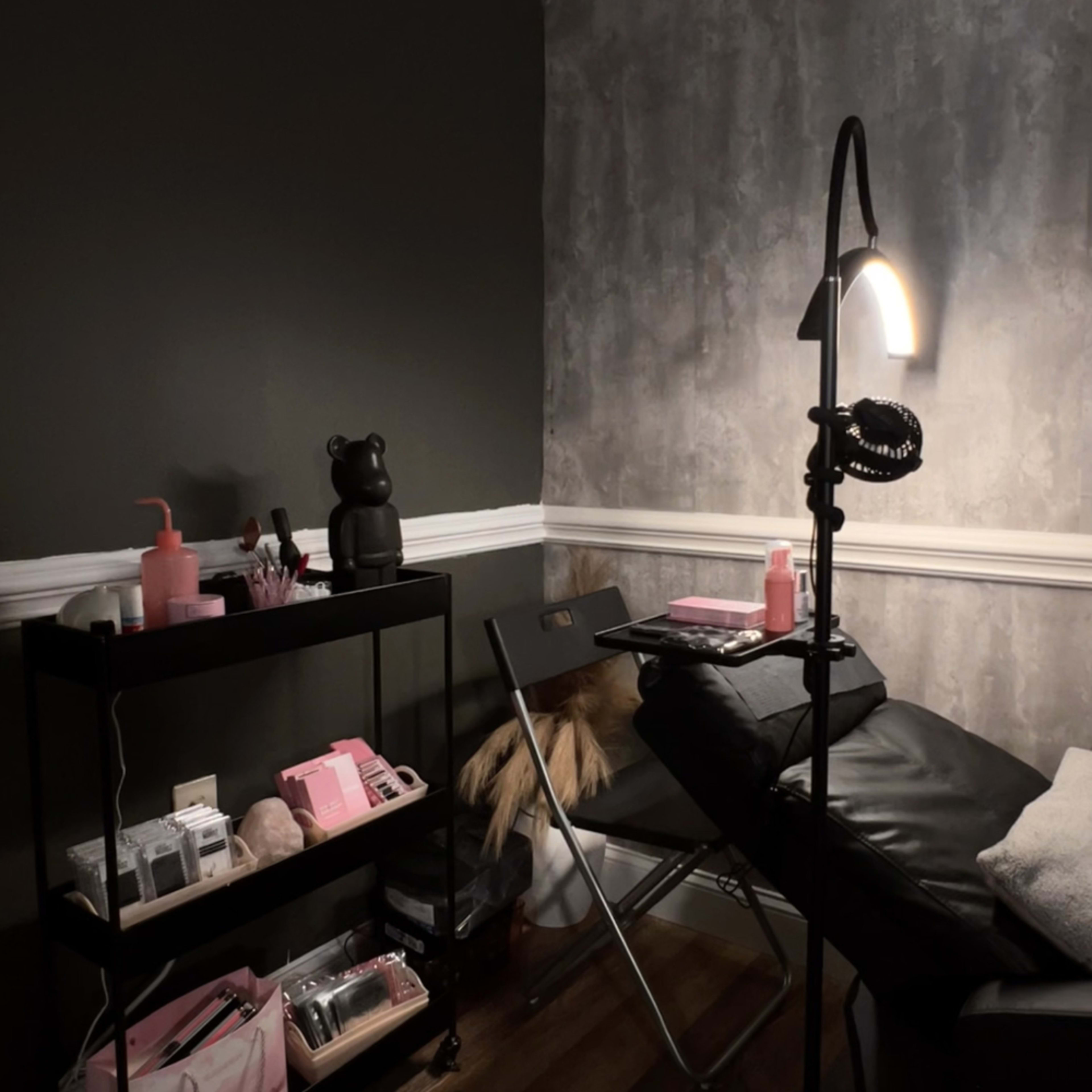 The image shows a dimly lit beauty room with a black treatment bed, a cart with various beauty products, and a standing lamp.