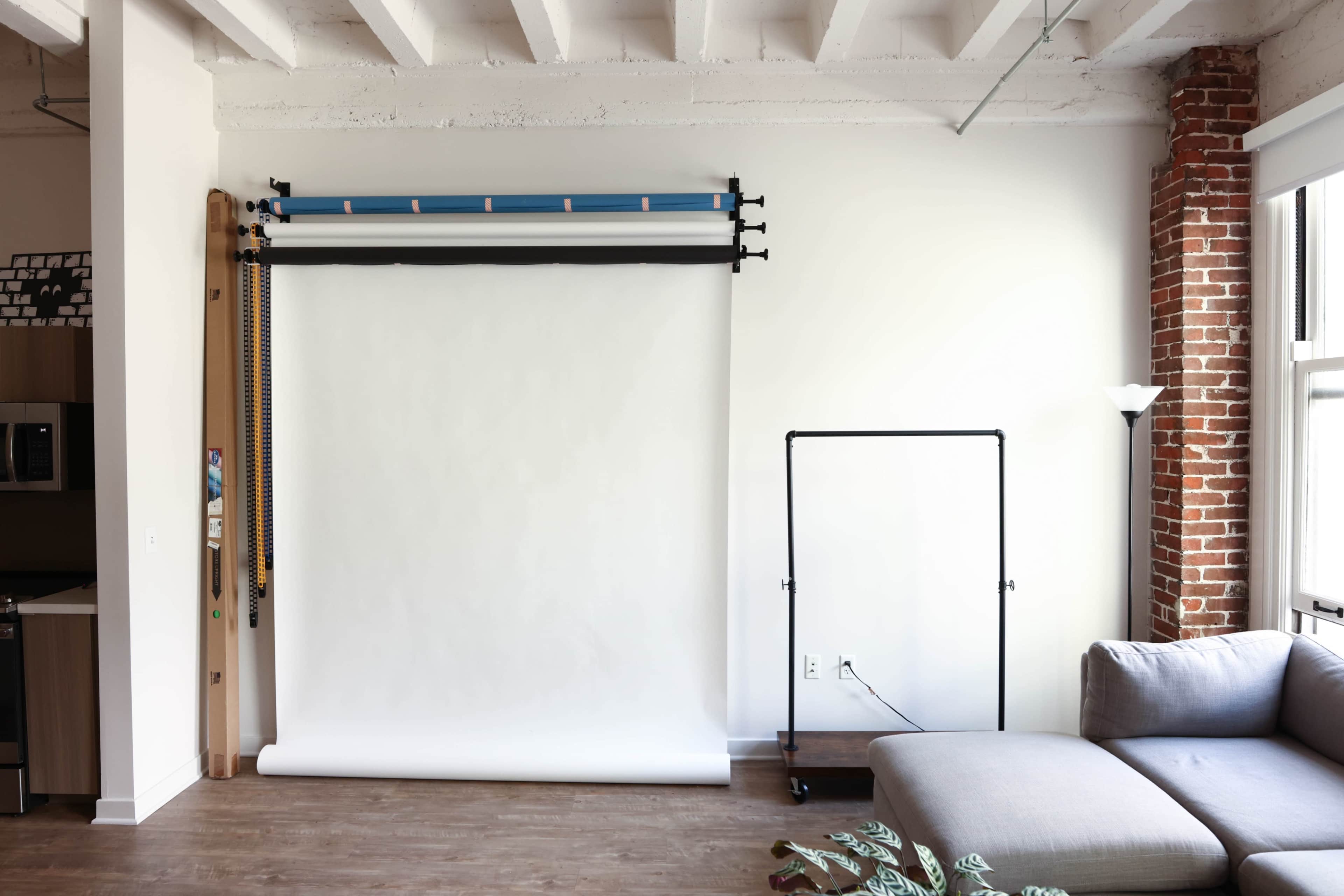 The image shows a studio space with a white backdrop mounted on the wall, a garment rack, and a grey sectional sofa, surrounded by natural light.