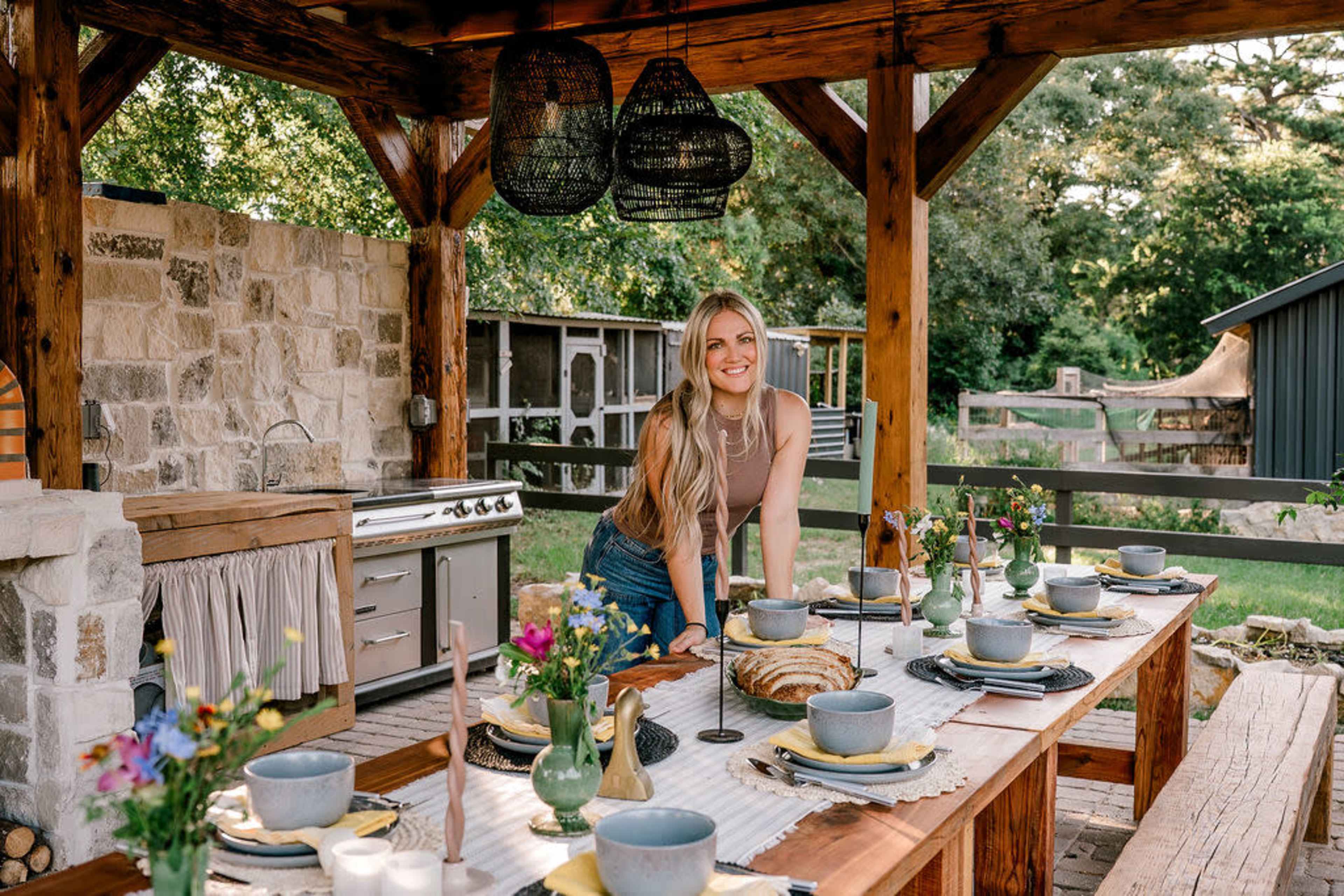 A woman sets a rustic outdoor dining table adorned with dishes and flowers under a wooden pergola.