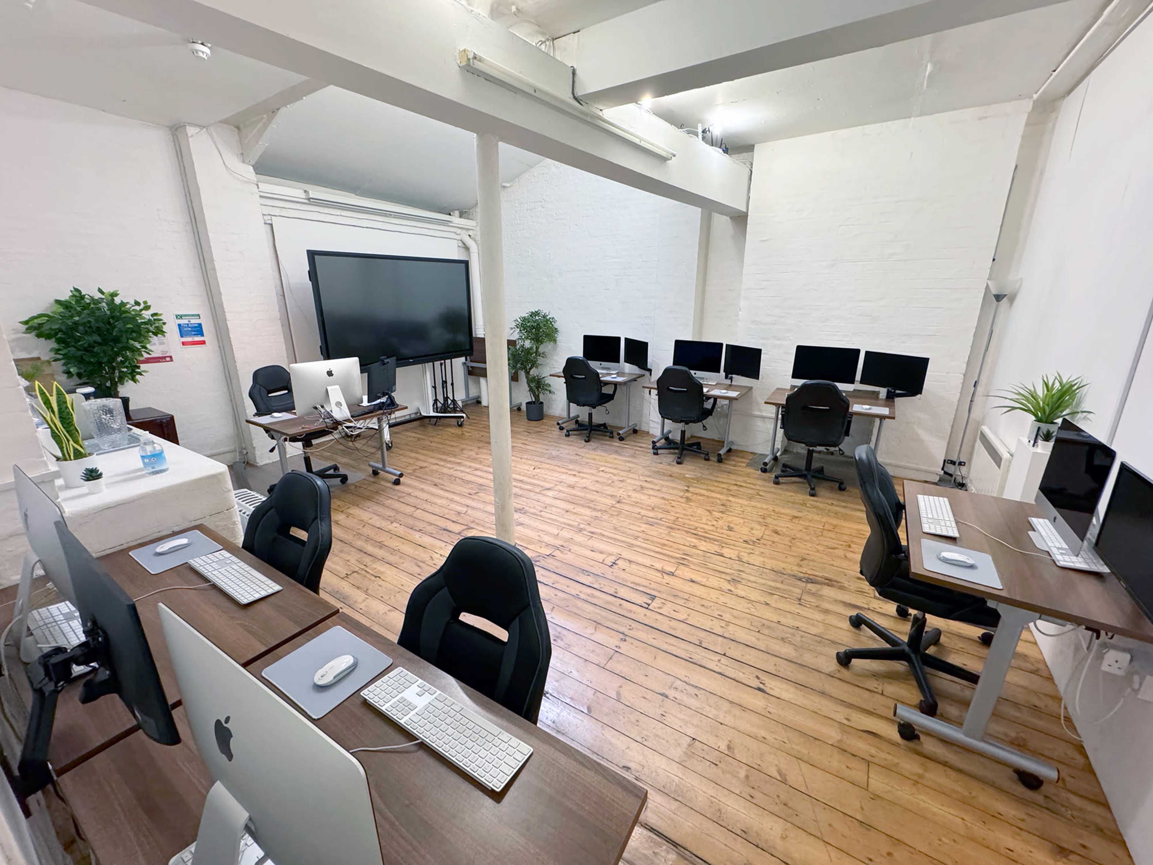 A spacious office with several desks equipped with Apple computers and ergonomic chairs, along with potted plants, arranged around a large display screen.
