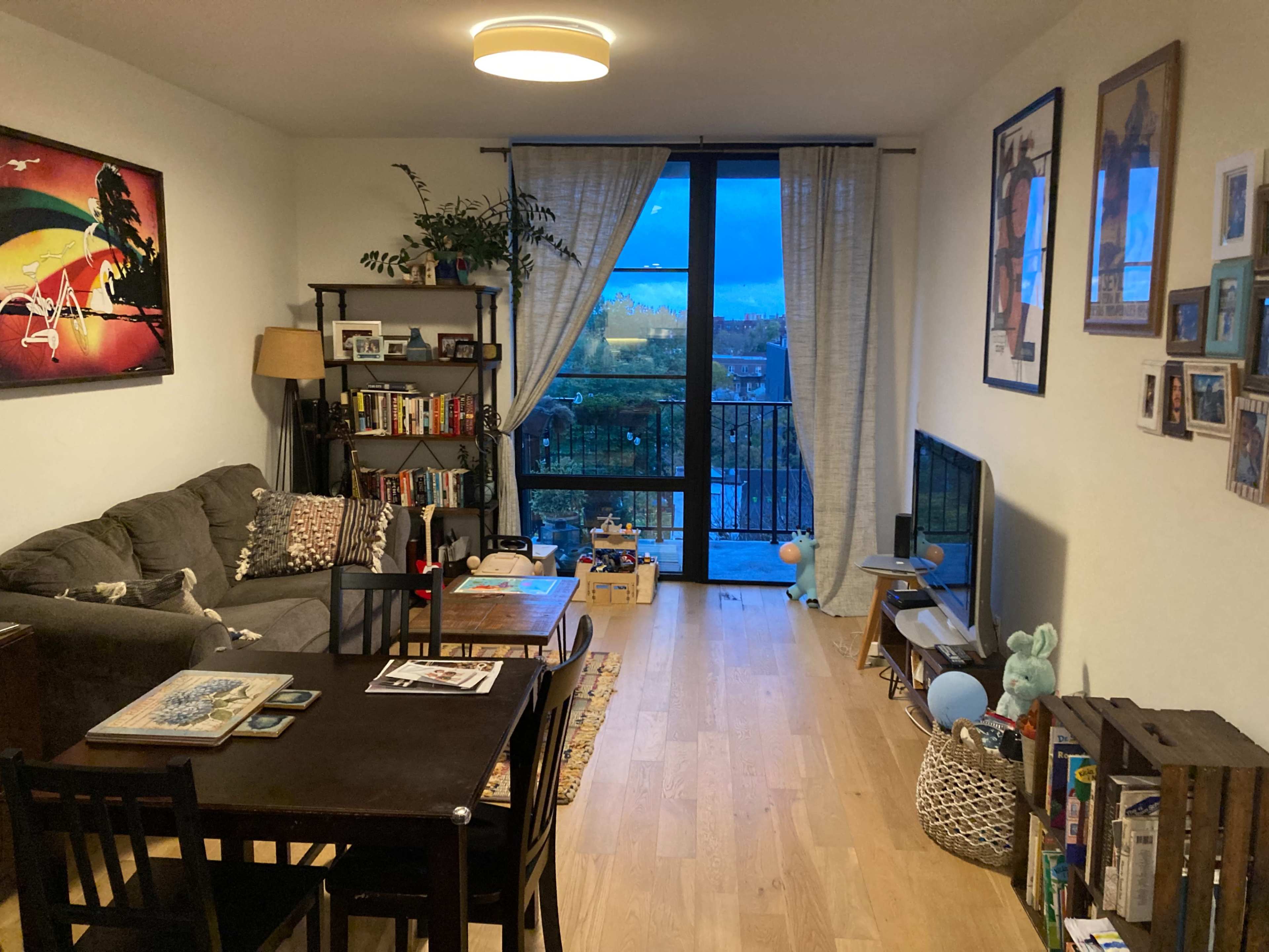 The image depicts a cozy living room with a gray sofa, a wooden dining table, shelves of books, and large windows overlooking a scenic view.