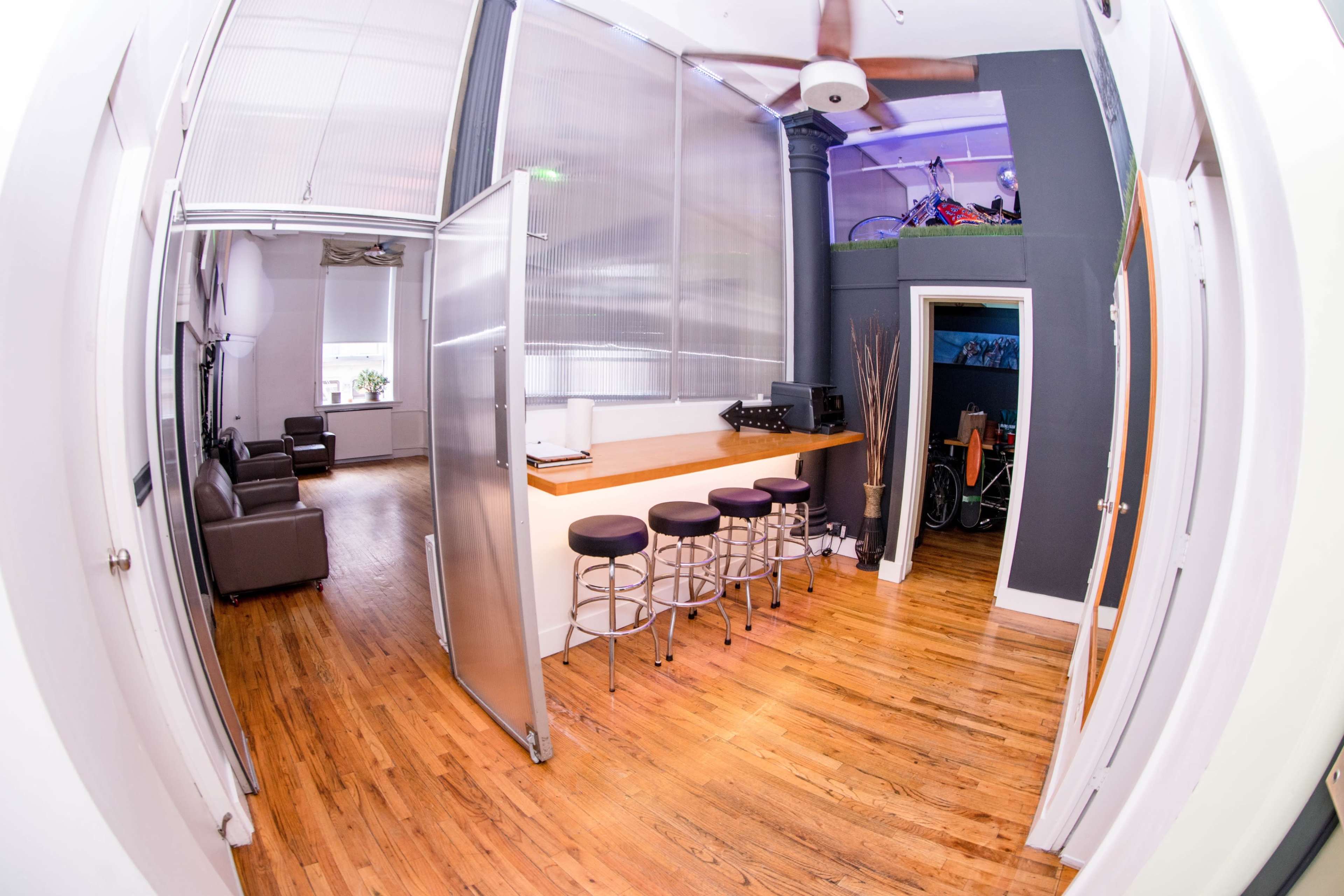 The image shows a spacious, modern office interior featuring a bar-like counter with stools, a seating area, and a room that includes a bicycle and a workspace.