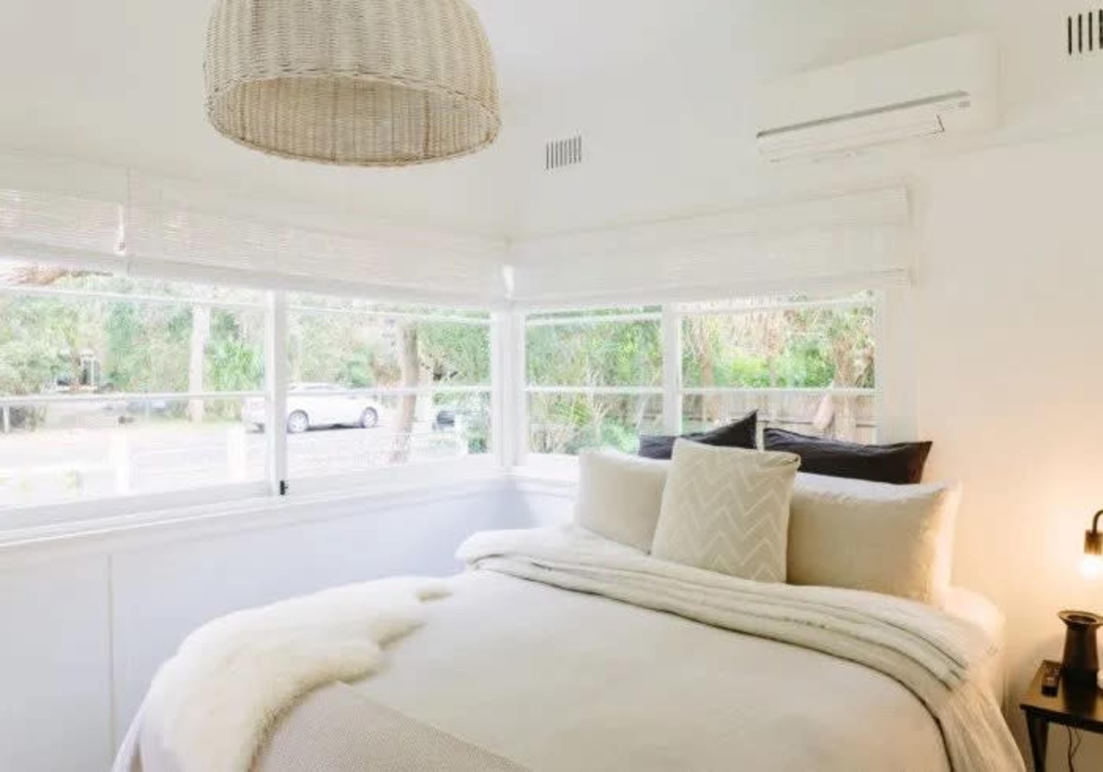 A bright bedroom with a bed covered in neutral bedding, large windows letting in natural light, and a woven pendant lamp hanging overhead.