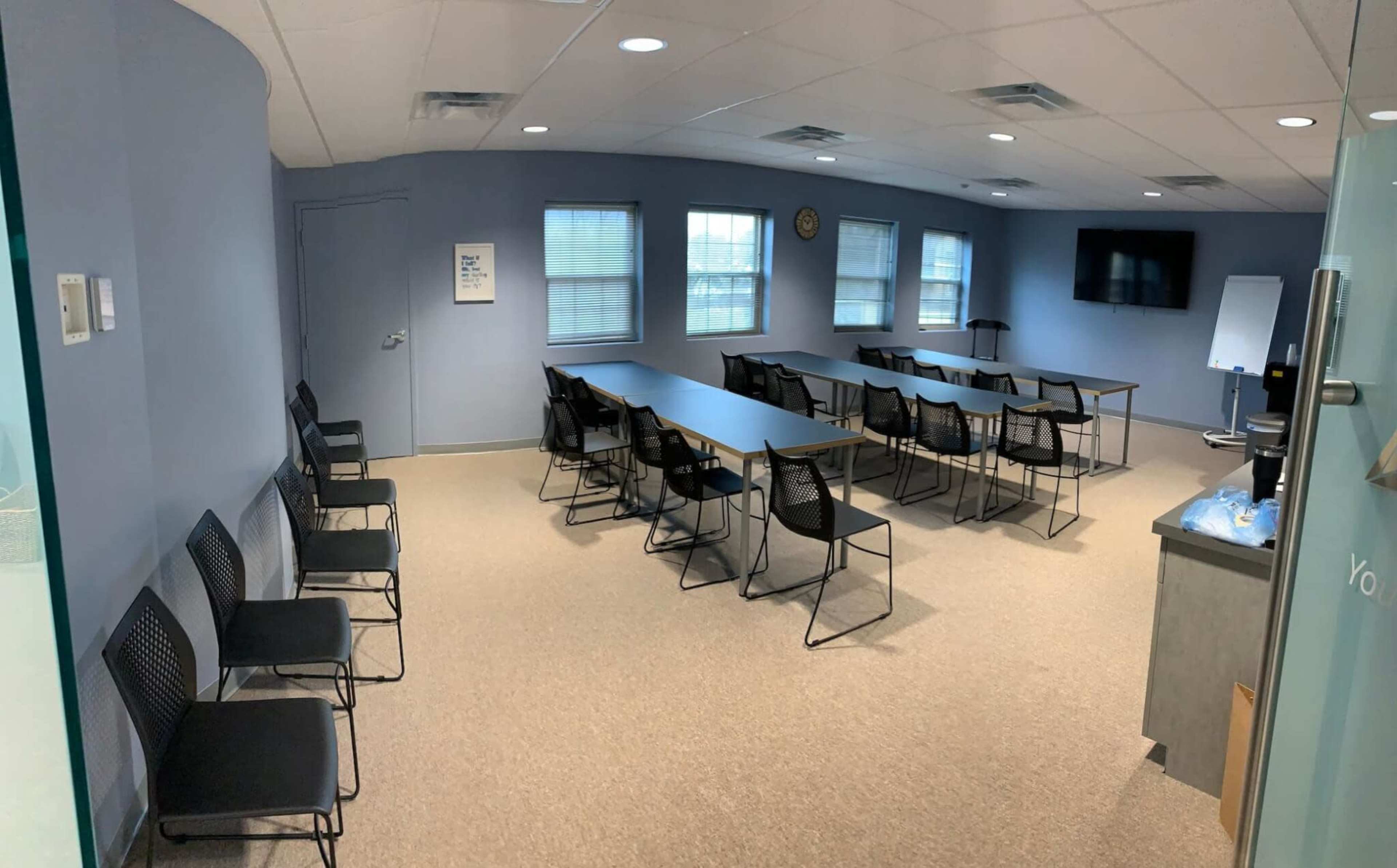 A conference room with multiple rows of chairs and long tables arranged for a meeting, featuring a television and a clock on the wall.