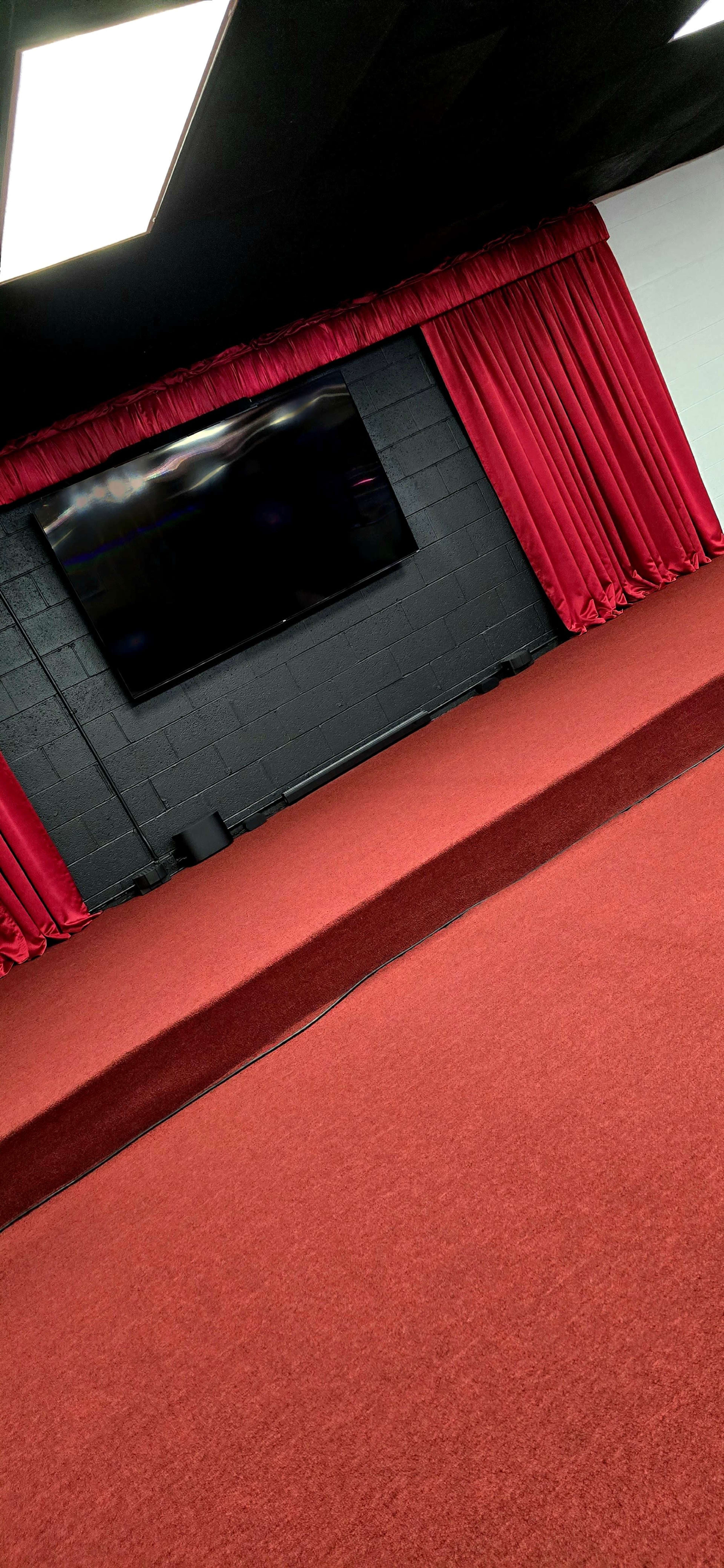 The image features a performance space with red carpet, red curtains, and a large black screen on a wall.