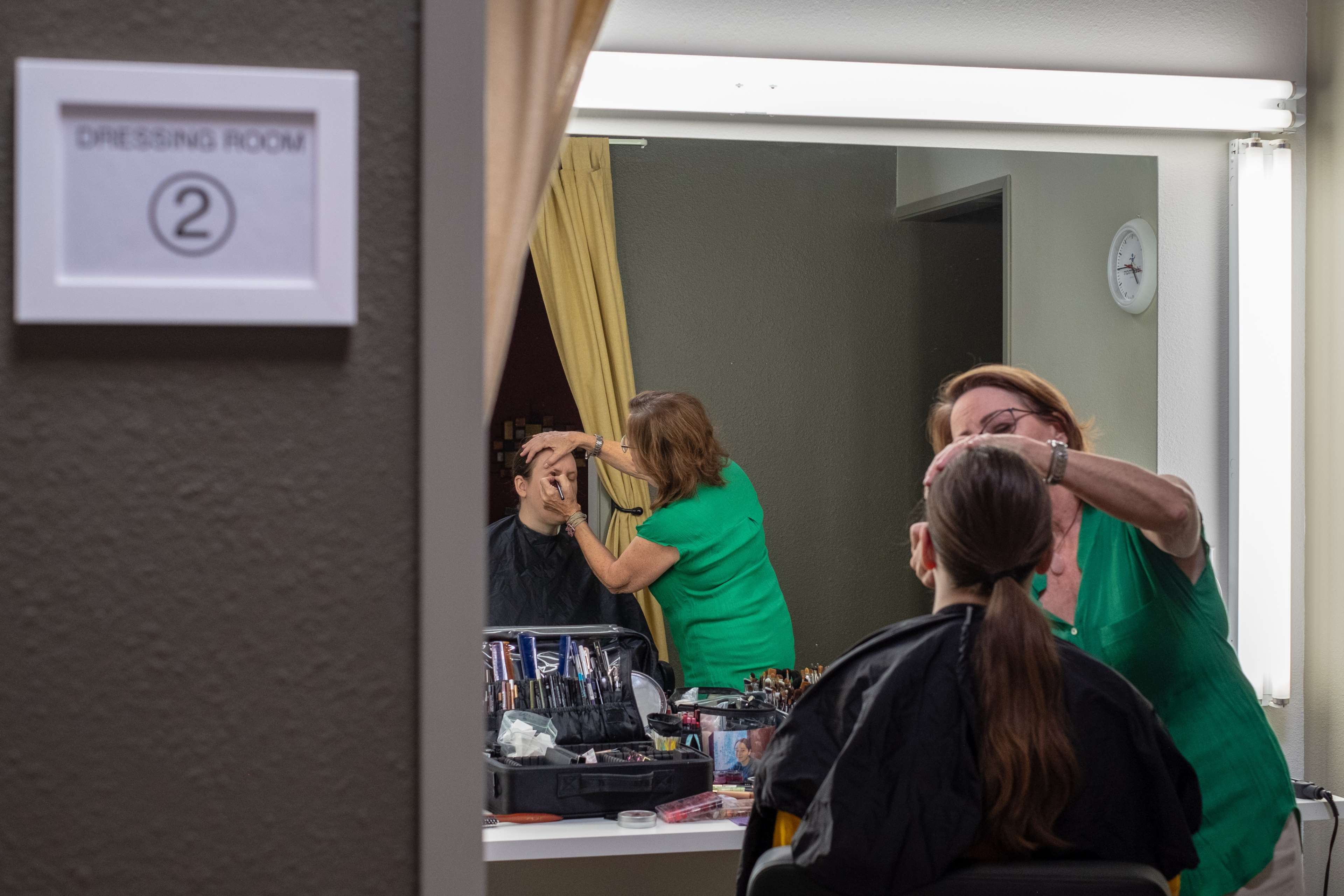 A makeup artist applies makeup to a client in a dressing room, reflecting the scene in a mirror.