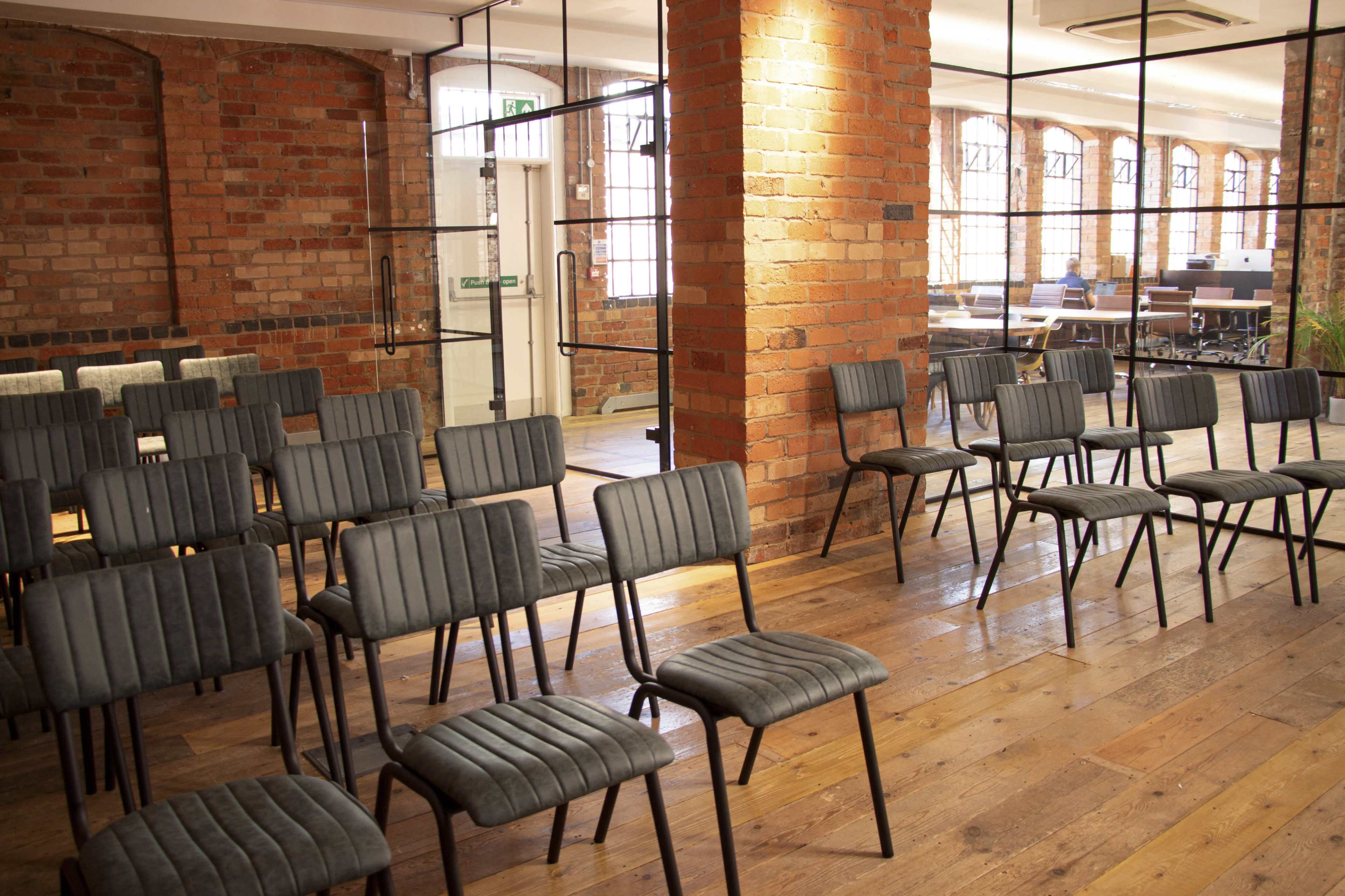 The image shows a room with rows of metal-framed chairs arranged on wooden flooring, with brick walls and large windows in the background.