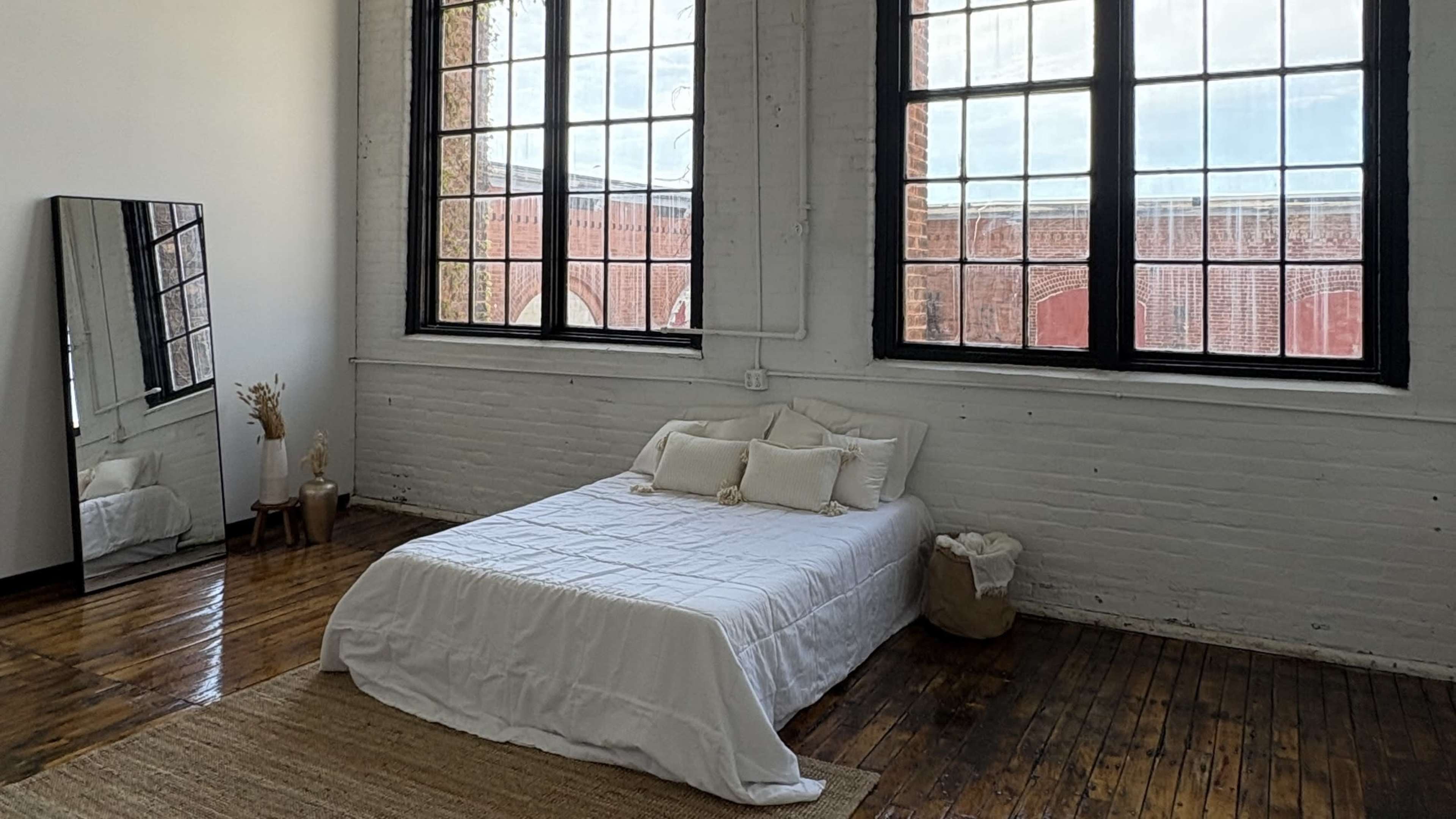 The room features a bed with a white coverlet, large windows letting in natural light, and a mirror against the wall.