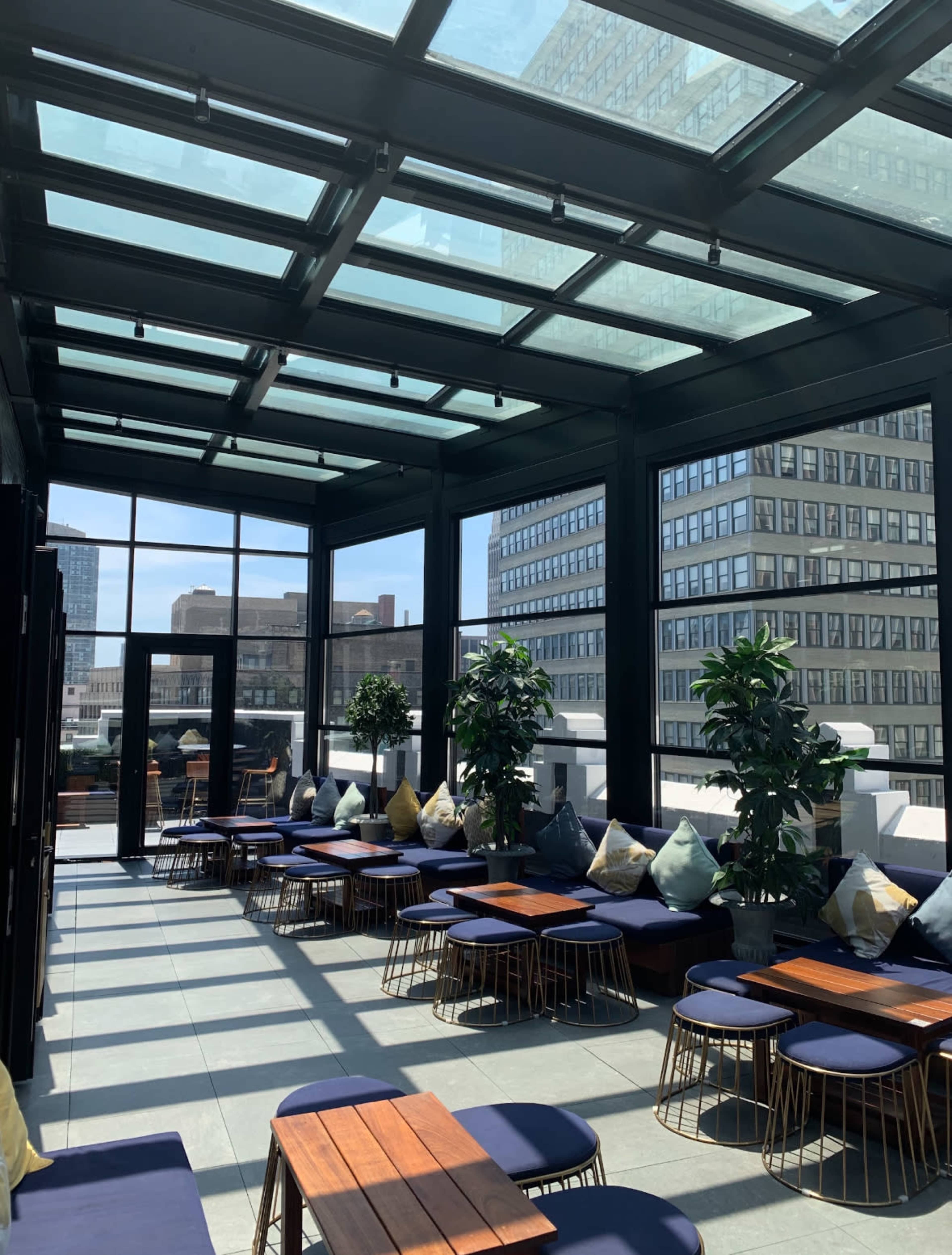 The image shows a rooftop lounge with glass ceilings, featuring low seating with dark cushions and wooden tables surrounded by potted plants.