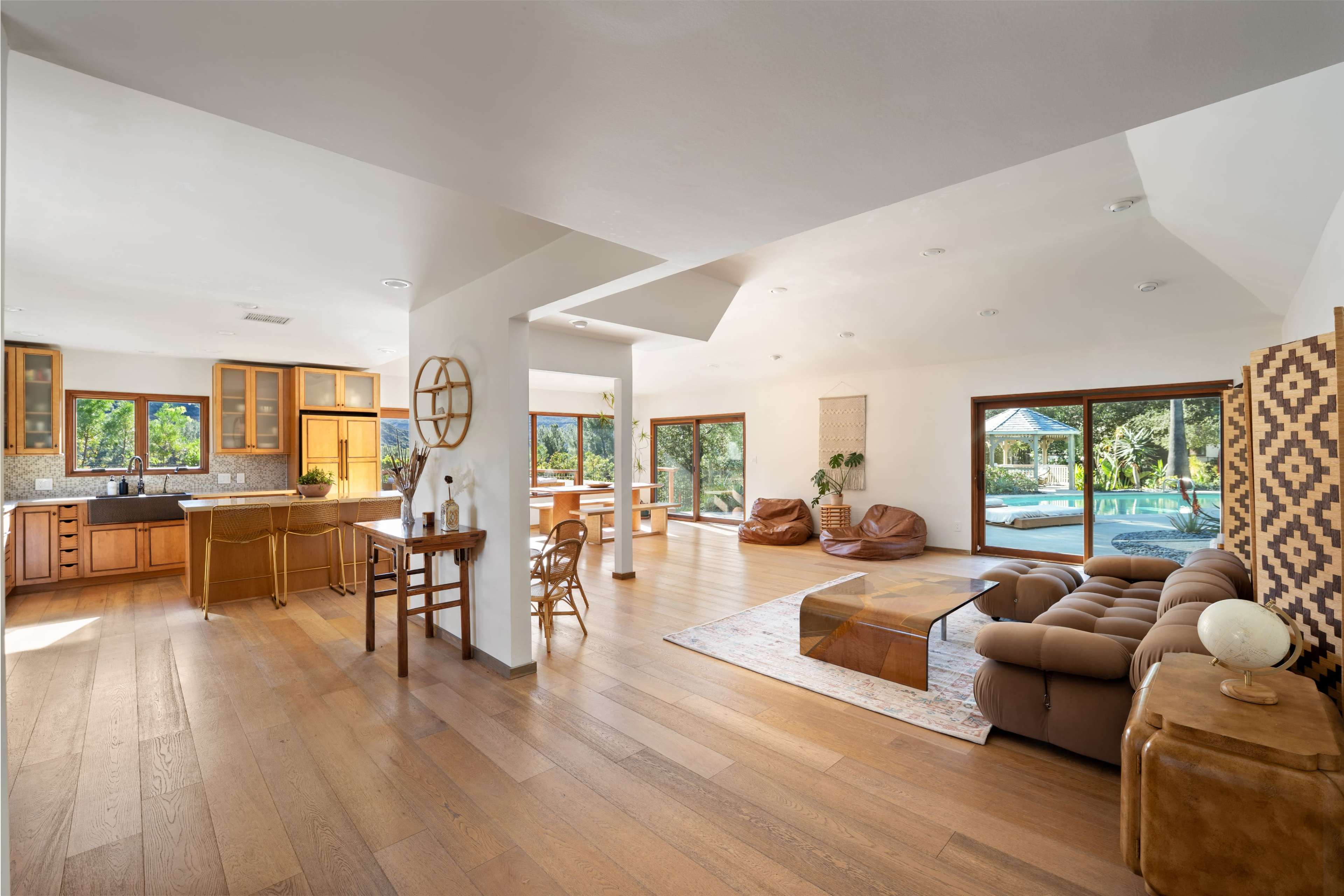 The image depicts a spacious, modern living area with a kitchen, dining space, and seating area that opens to a poolside view.