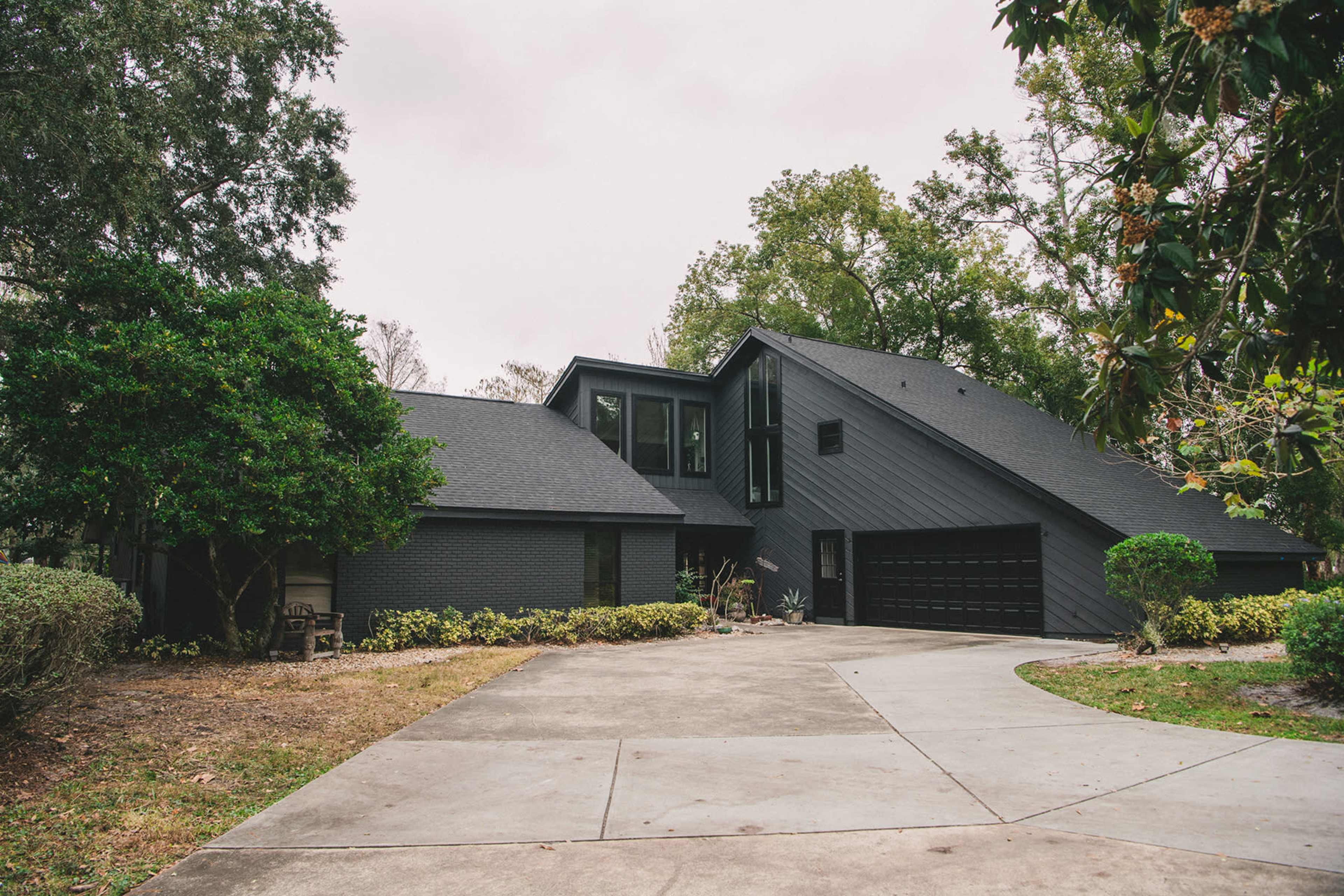 The image shows a modern, dark-colored house with large windows and a driveway surrounded by greenery.