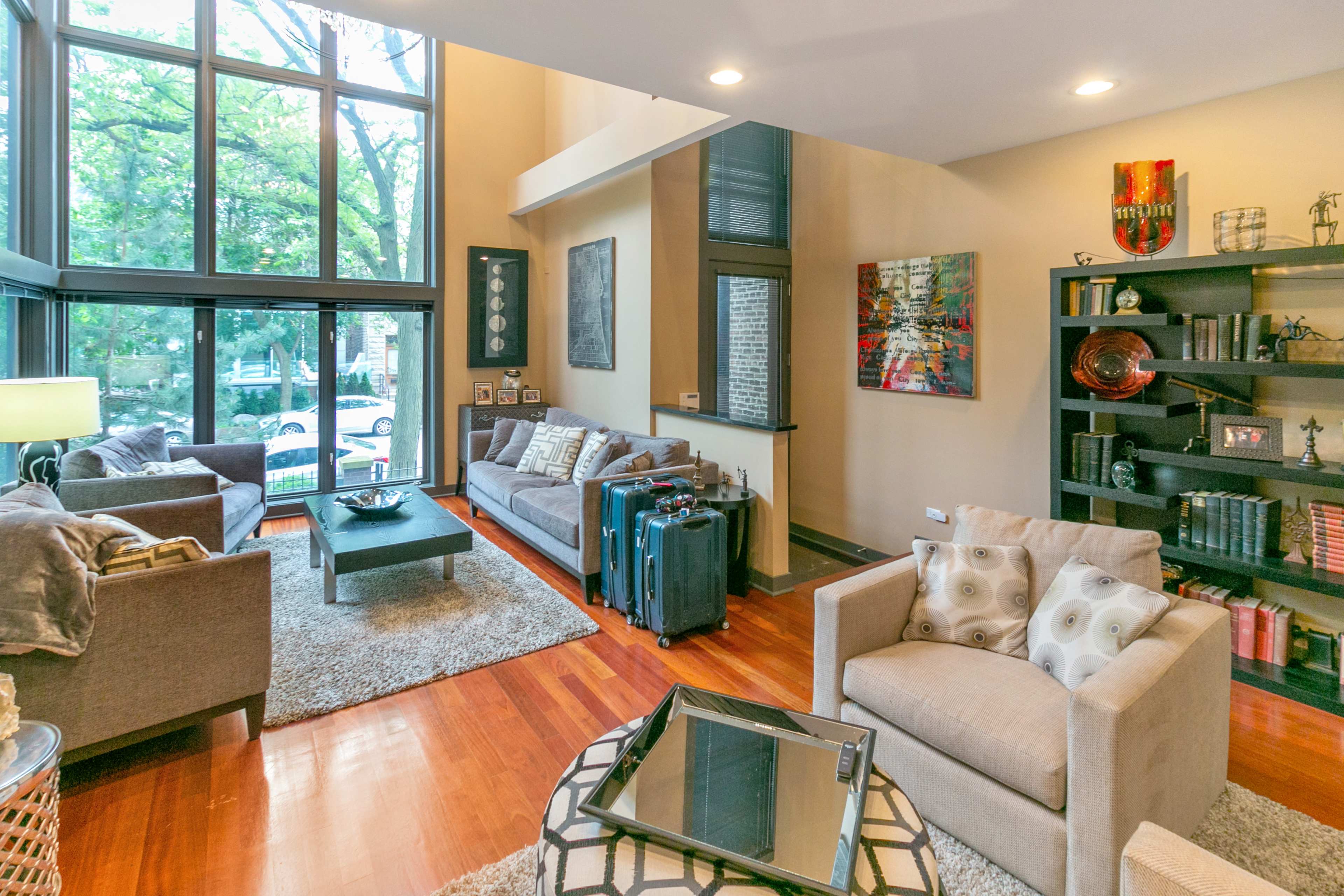 A modern living room features large windows, two sofas, an armchair, and a coffee table on a hardwood floor, with artwork and luggage visible.