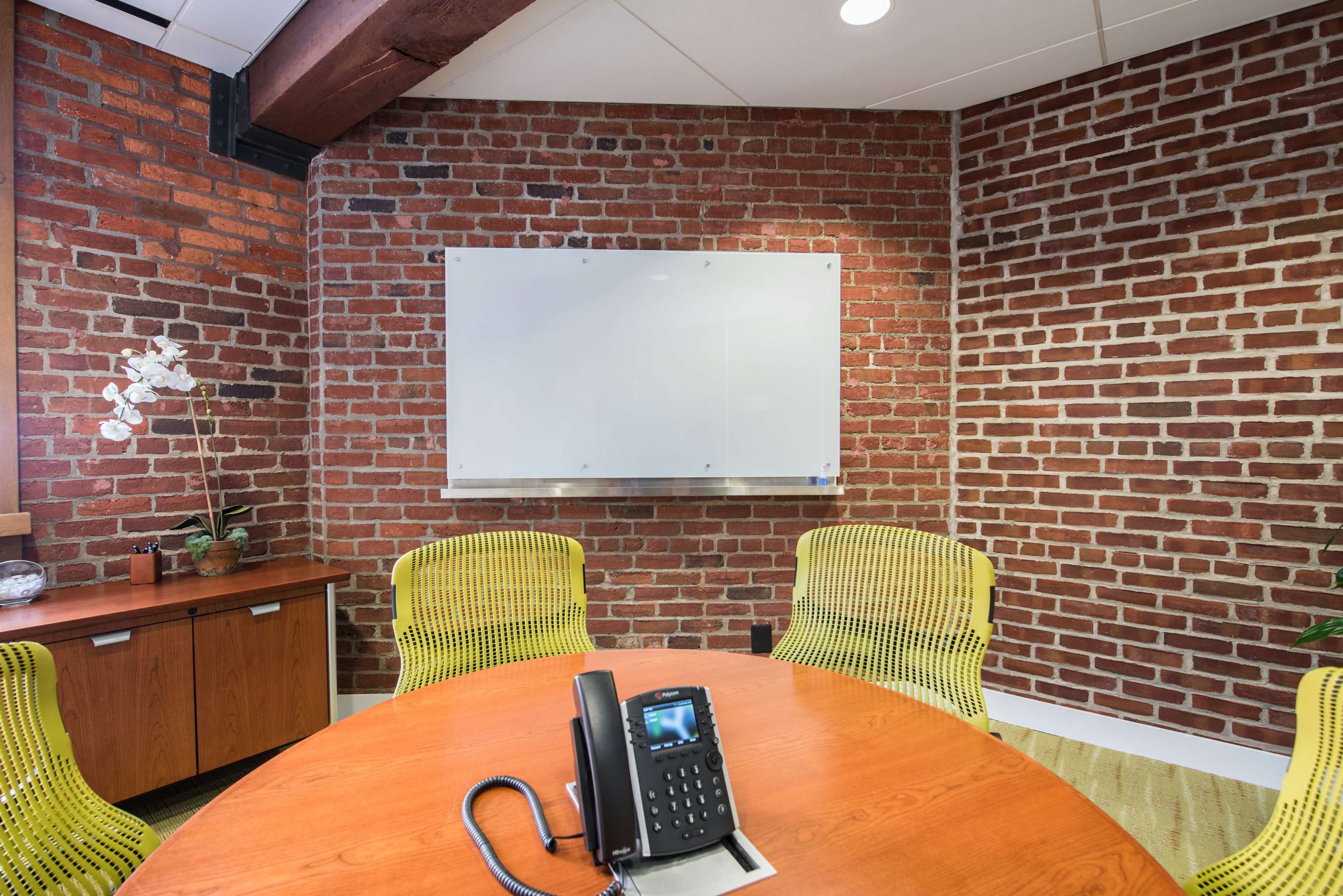 A conference room features a round wooden table with a phone, surrounded by four yellow chairs, and a whiteboard mounted on a brick wall.