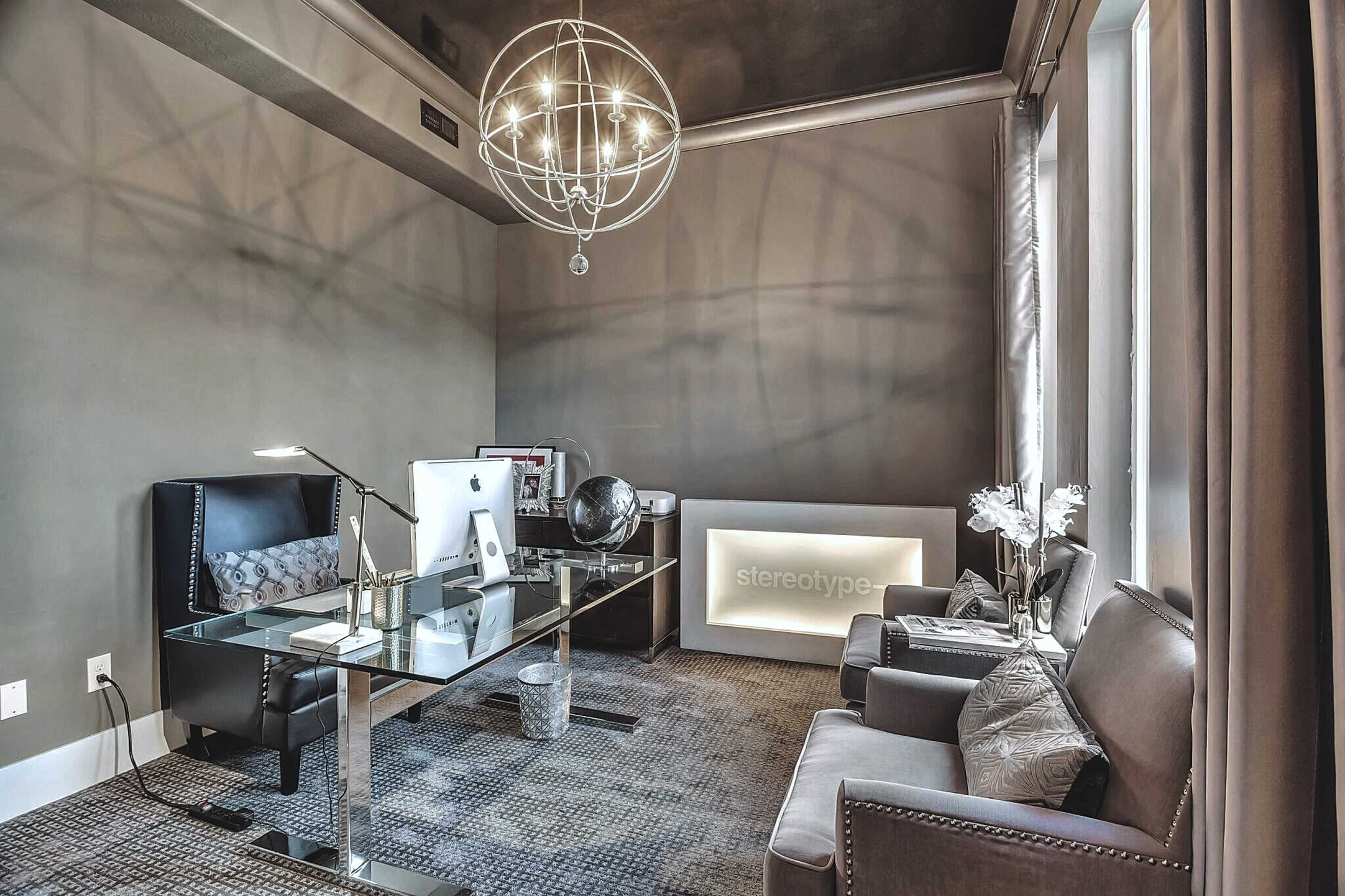 A modern office space featuring a glass desk with a computer, a black leather chair, a gray sofa, and decorative elements including a globe and a chandelier.