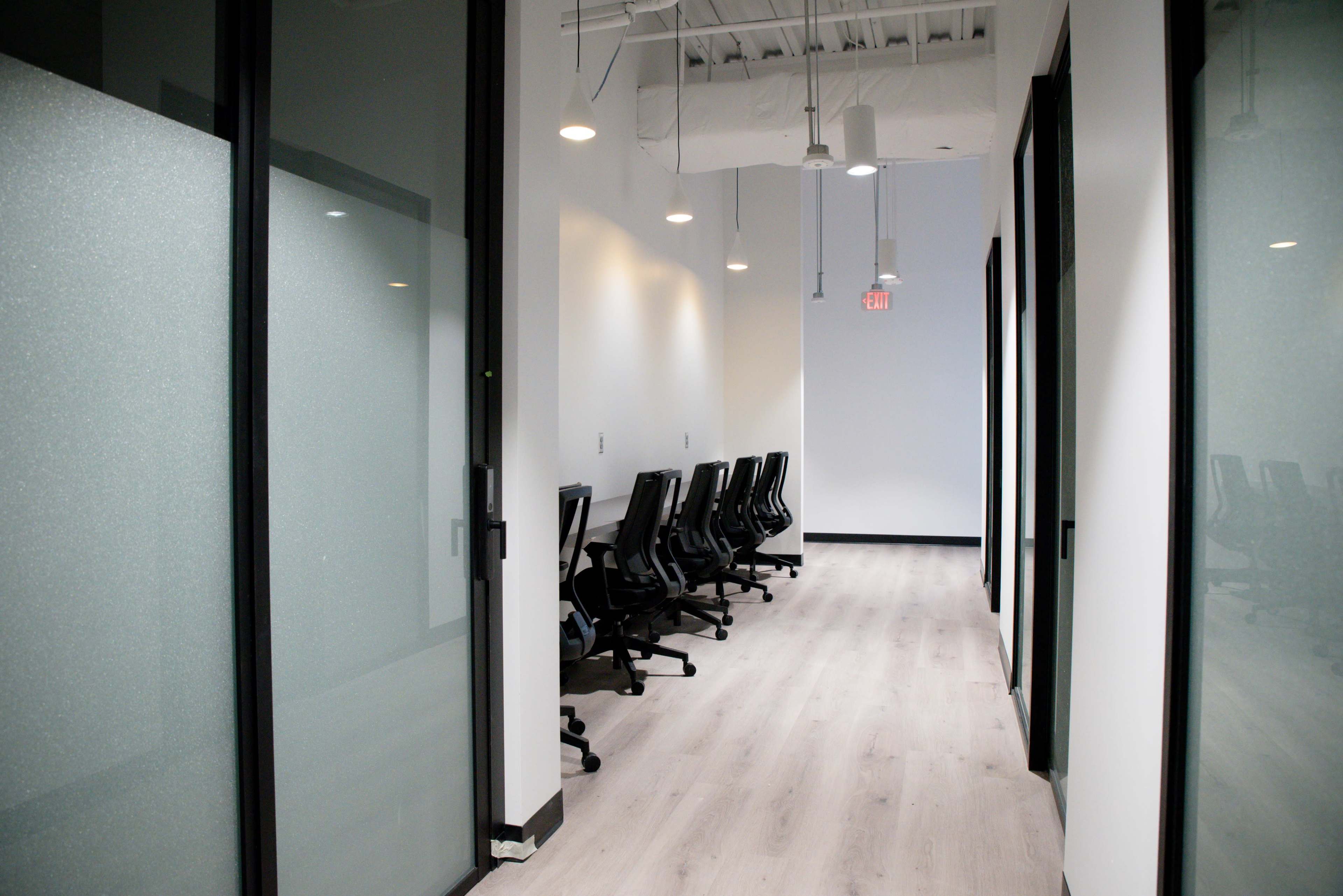 The image shows a modern office corridor with several chairs lined up against the wall and glass partitions on either side.