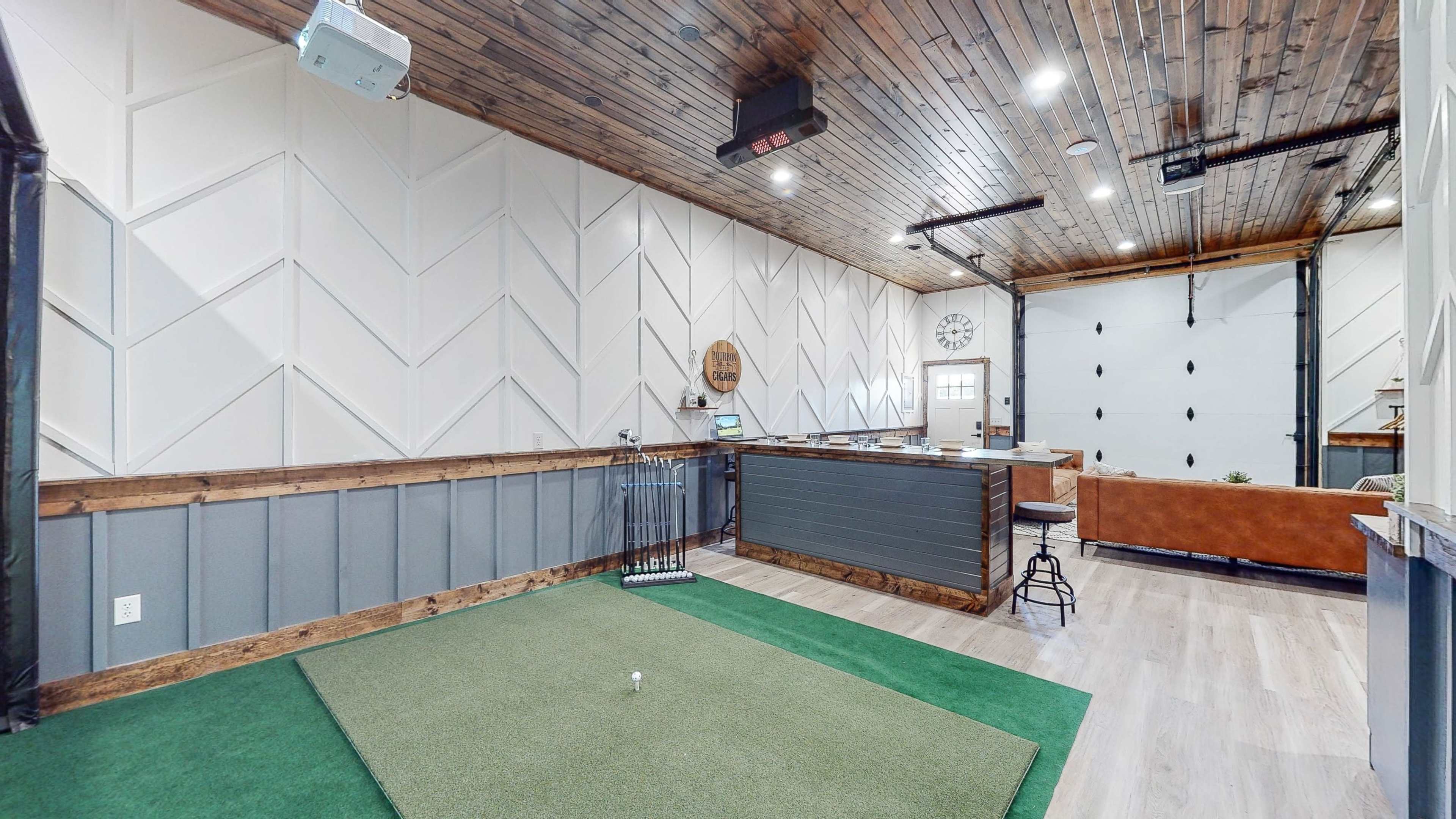 A spacious room with a putting green in the foreground, a bar area against one wall, and a cozy seating area visible in the background.