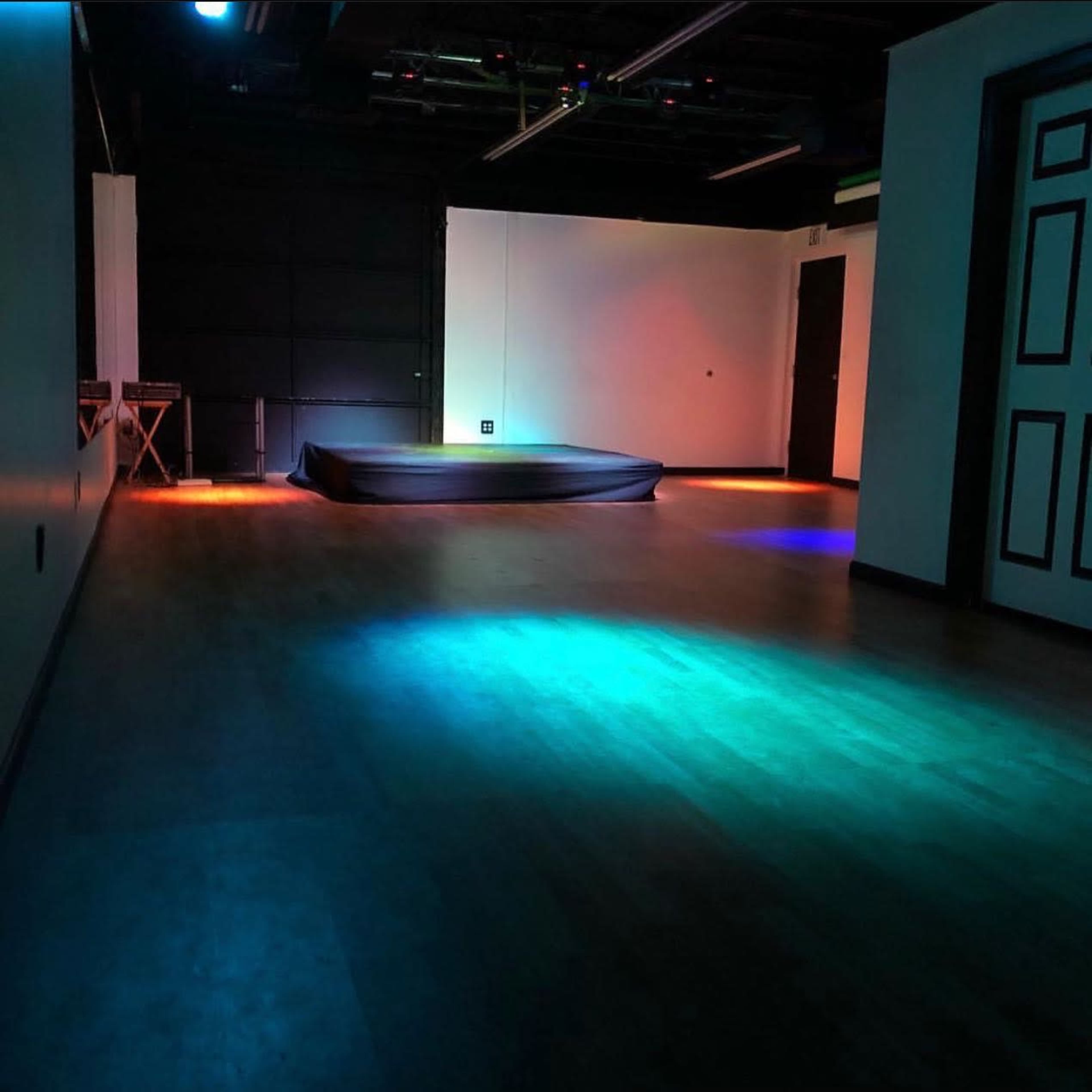 The image shows an empty room with a dark floor, a raised platform in the center, and colored lights illuminating various areas of the floor.