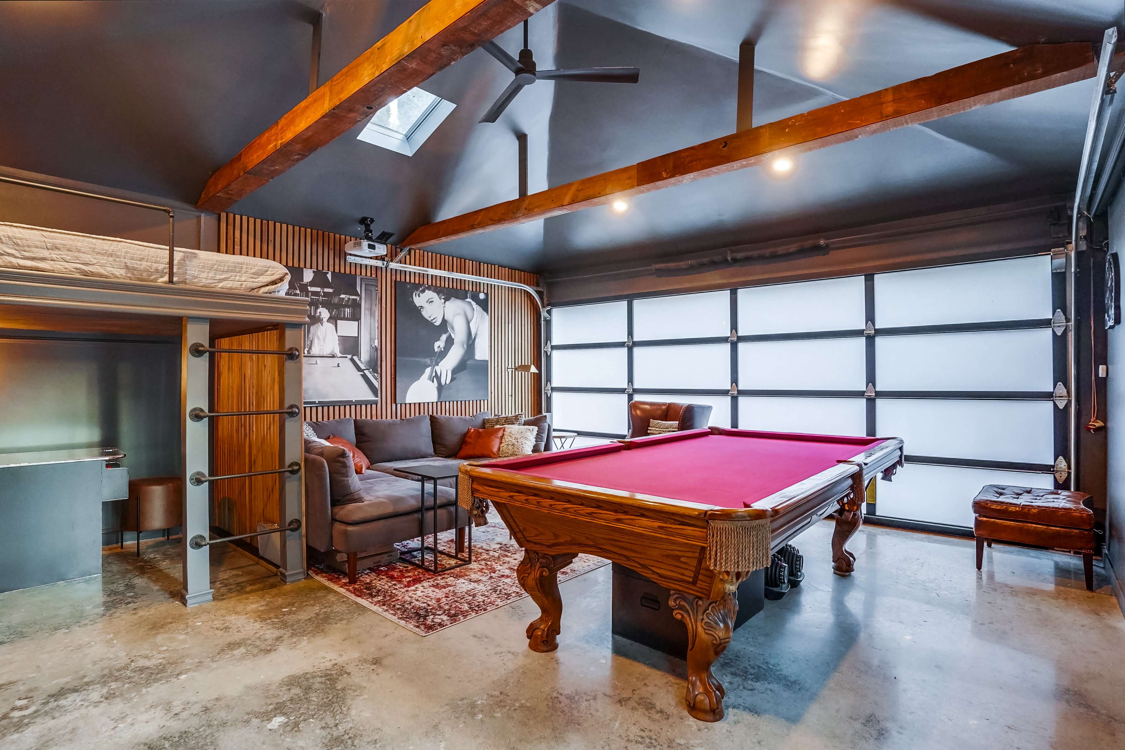 A modern game room featuring a billiards table, a seating area, and a large garage door, with wooden beams and a photo on the wall.