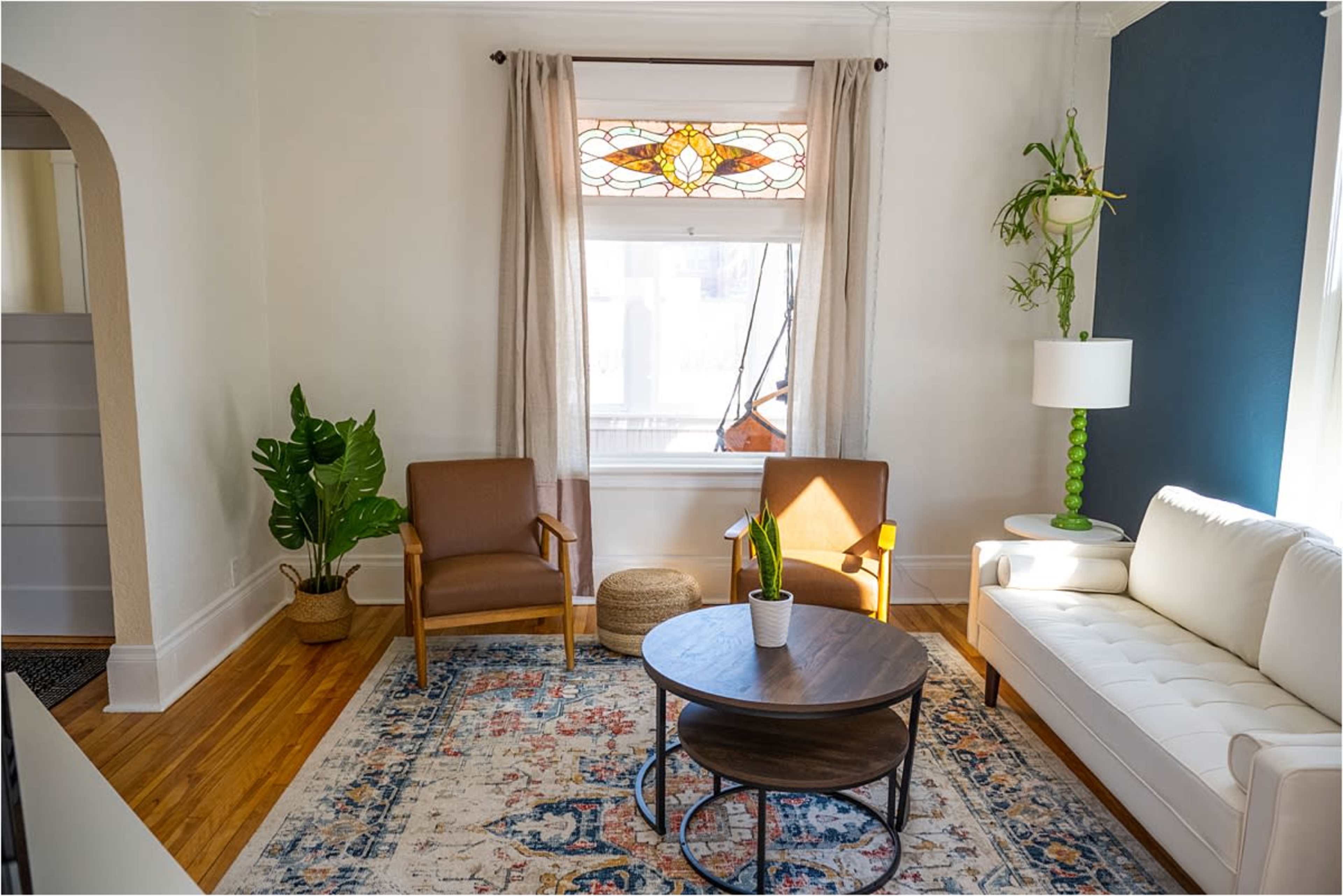 A bright living room featuring a white couch, two brown chairs, a round coffee table, a potted plant, and a lamp beside a window with a stained glass design.