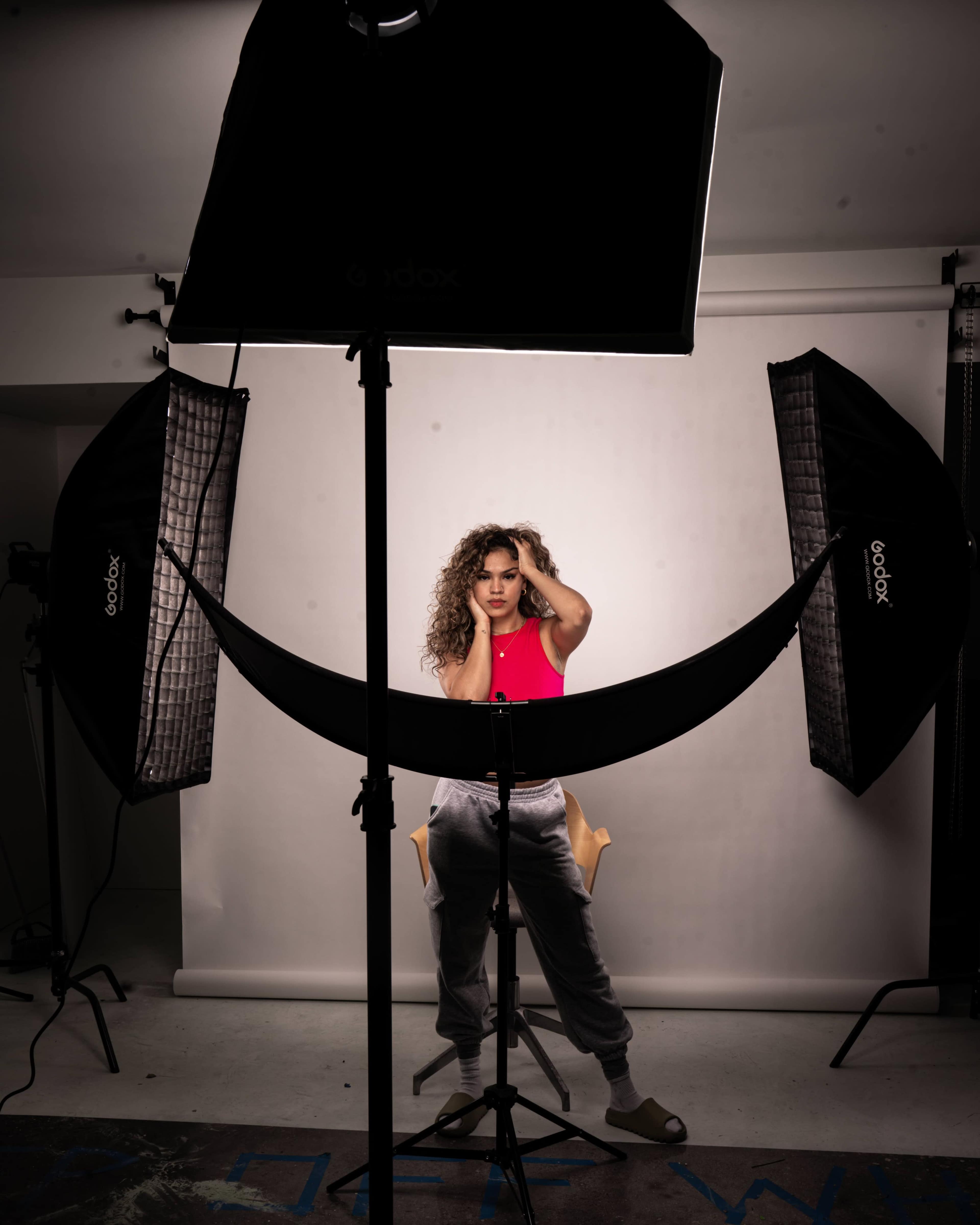 A person poses in a photography studio, surrounded by softbox lights and sitting on a stool against a white backdrop.