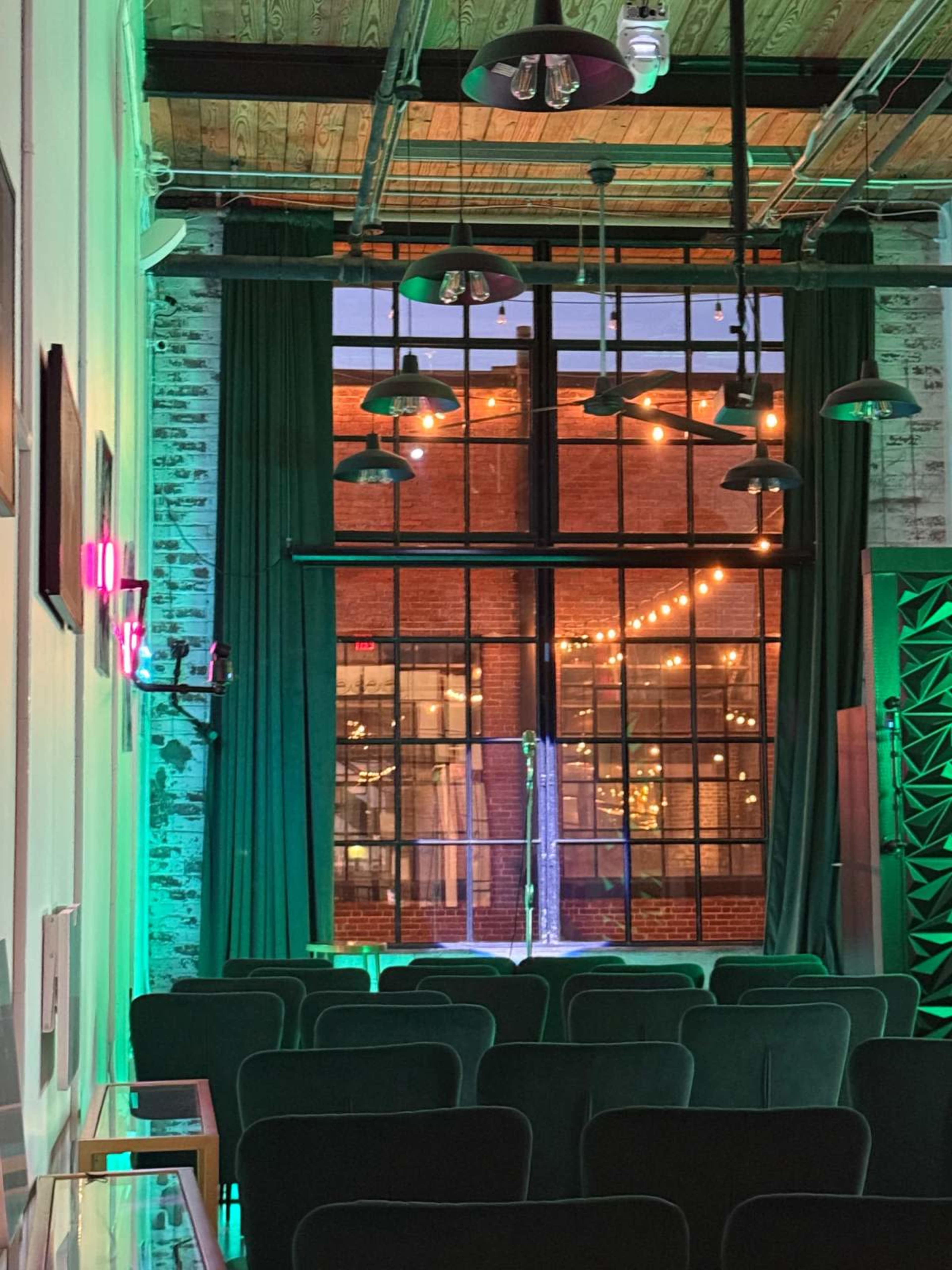 The image shows an indoor venue with green and pink lighting, featuring a row of empty seats facing a stage and large windows that reveal an evening outdoor view.