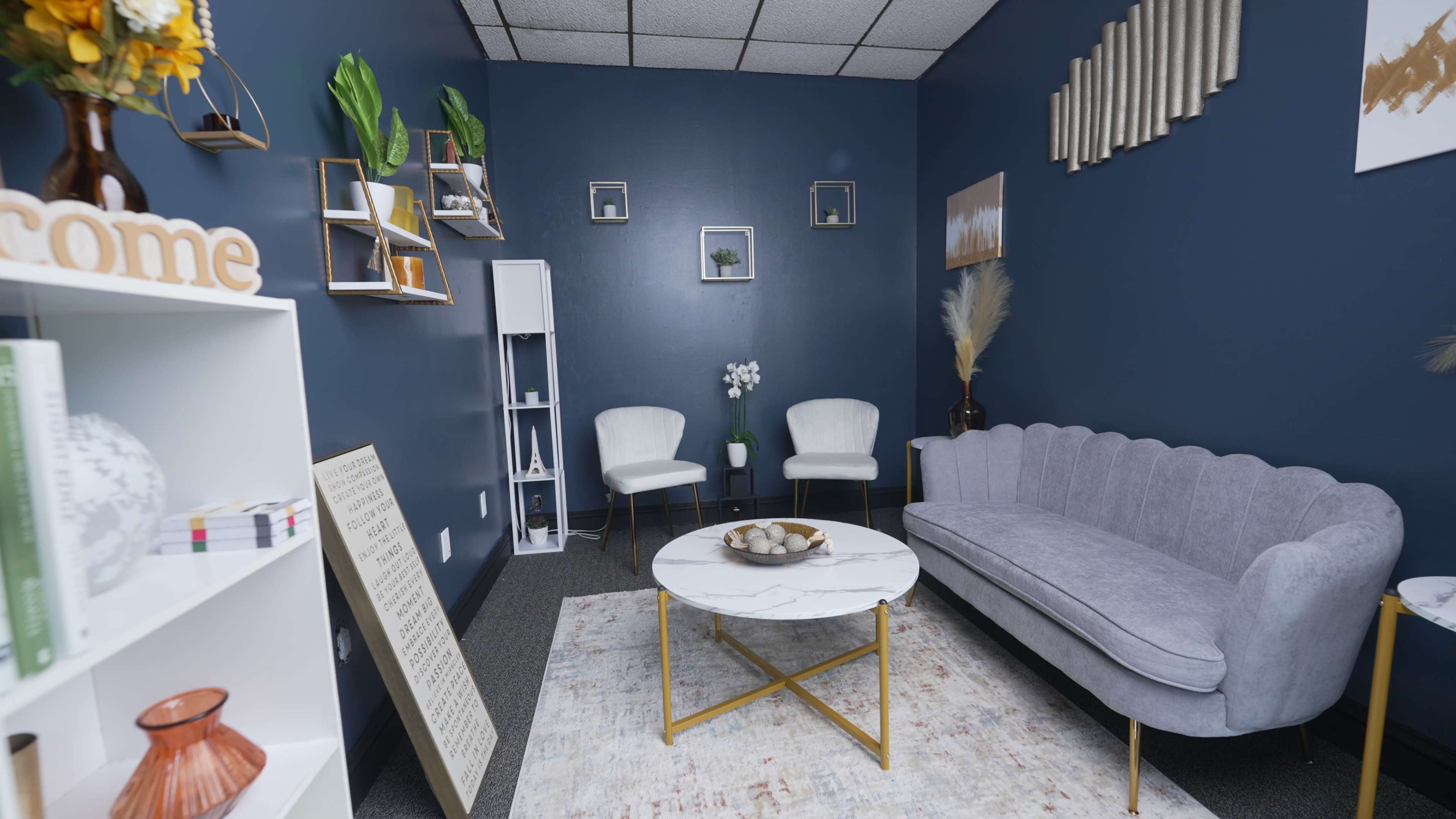 The image shows a cozy sitting area with a gray couch, two white chairs, a marble coffee table, and decorative shelves against a dark blue wall.