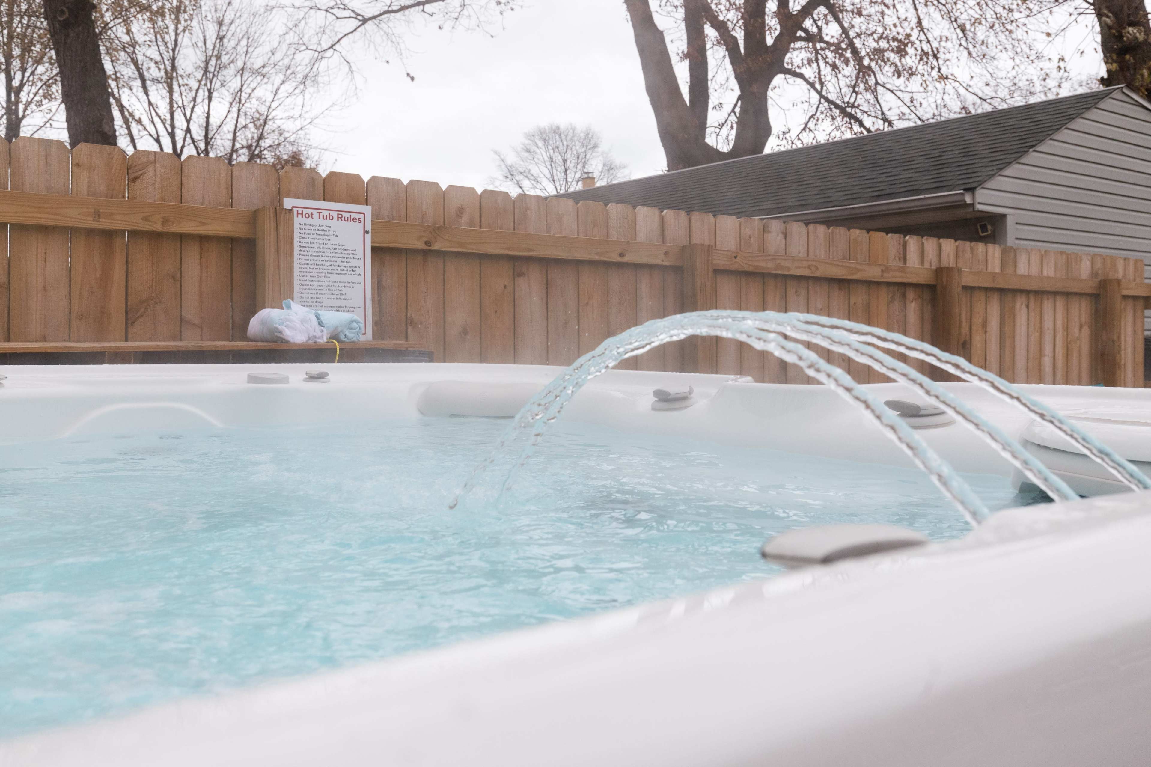 A hot tub with water jets creating arcs of water is located near a wooden fence, with a warning sign visible in the background.