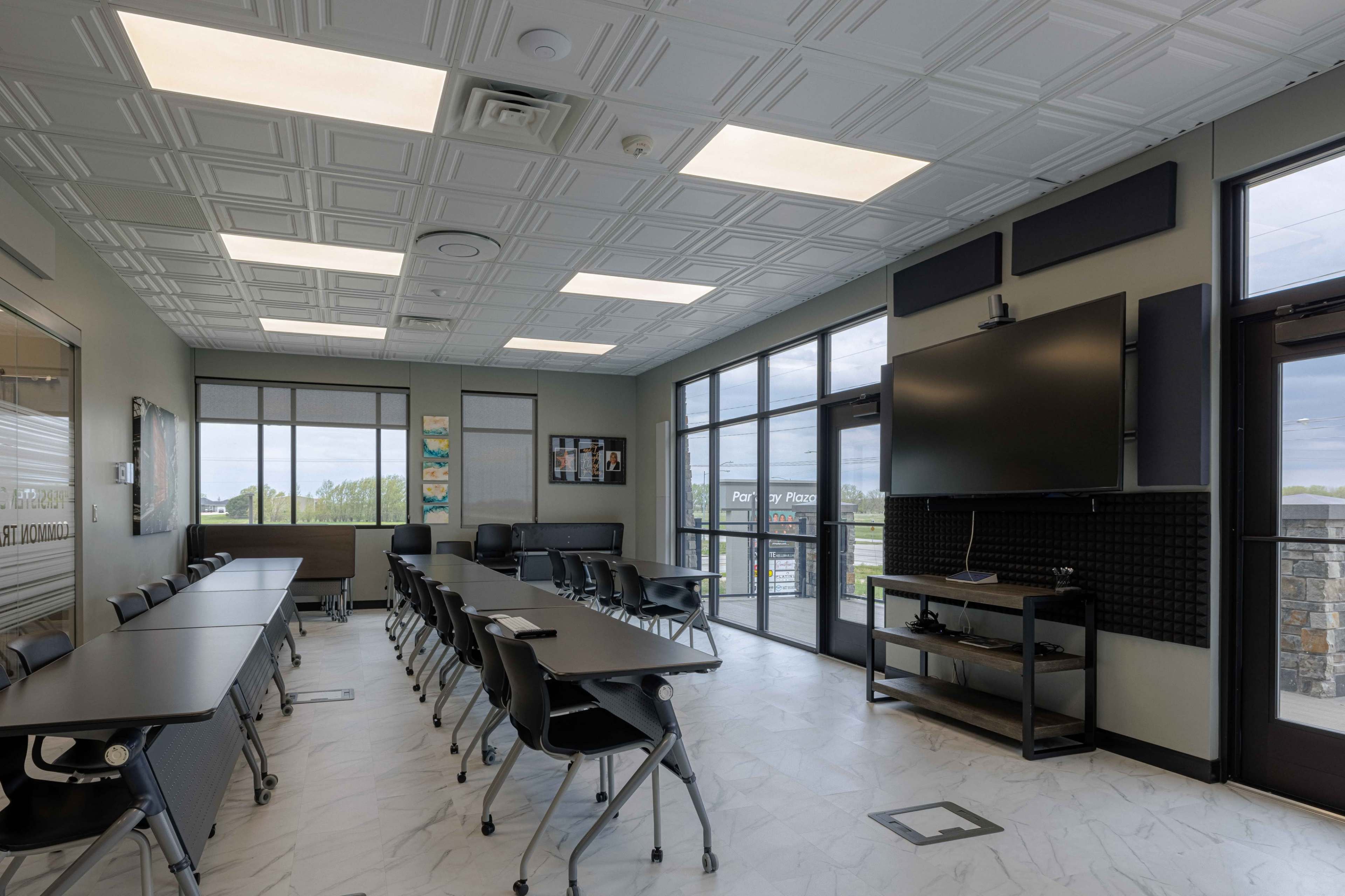 The image shows a modern classroom equipped with a large wall-mounted television, arranged desks, and windows overlooking an outdoor area.