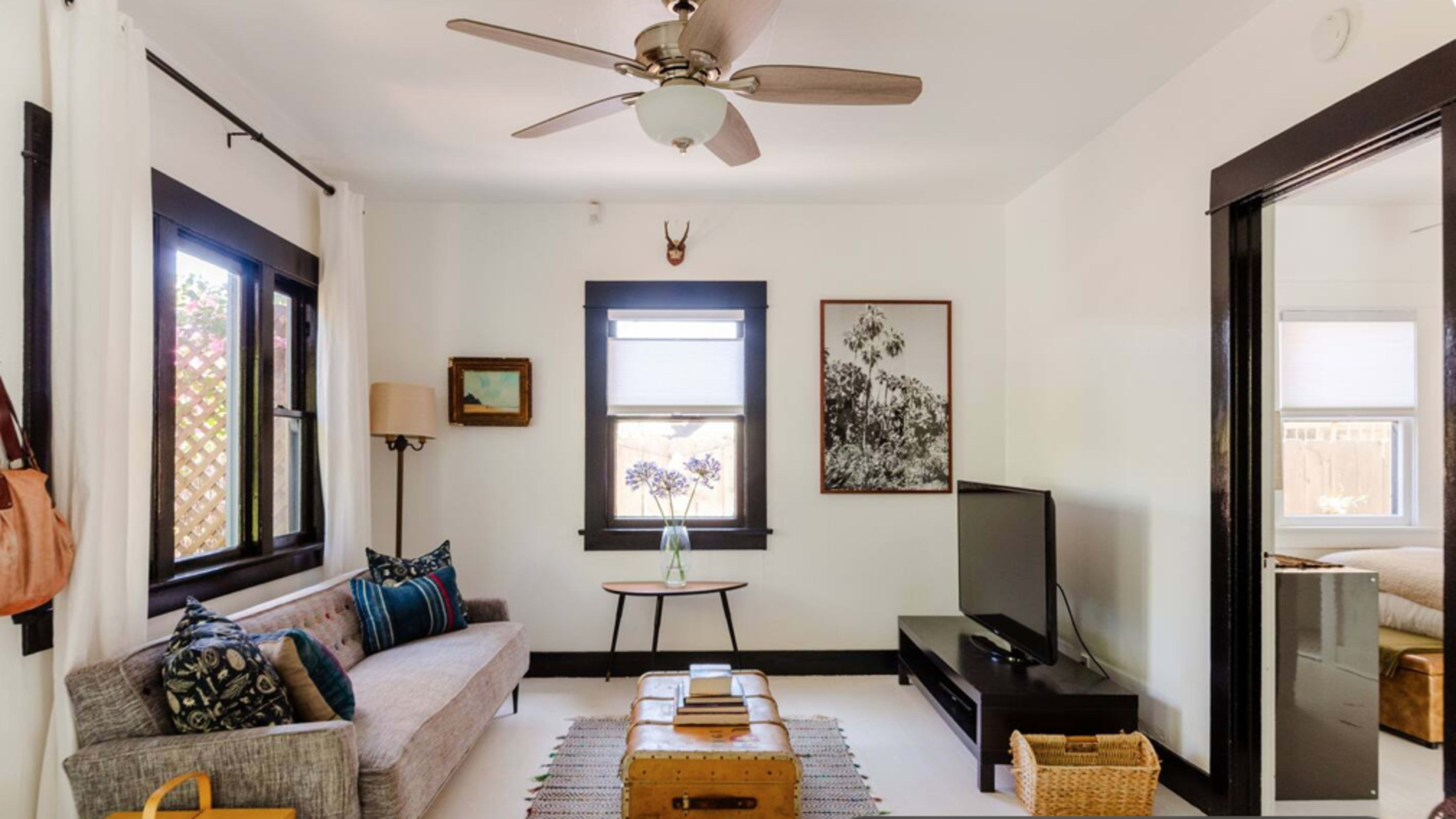 The image shows a cozy living room with a gray sofa, a wooden coffee table, a television, and several framed artworks on the walls, illuminated by natural light coming through the windows.