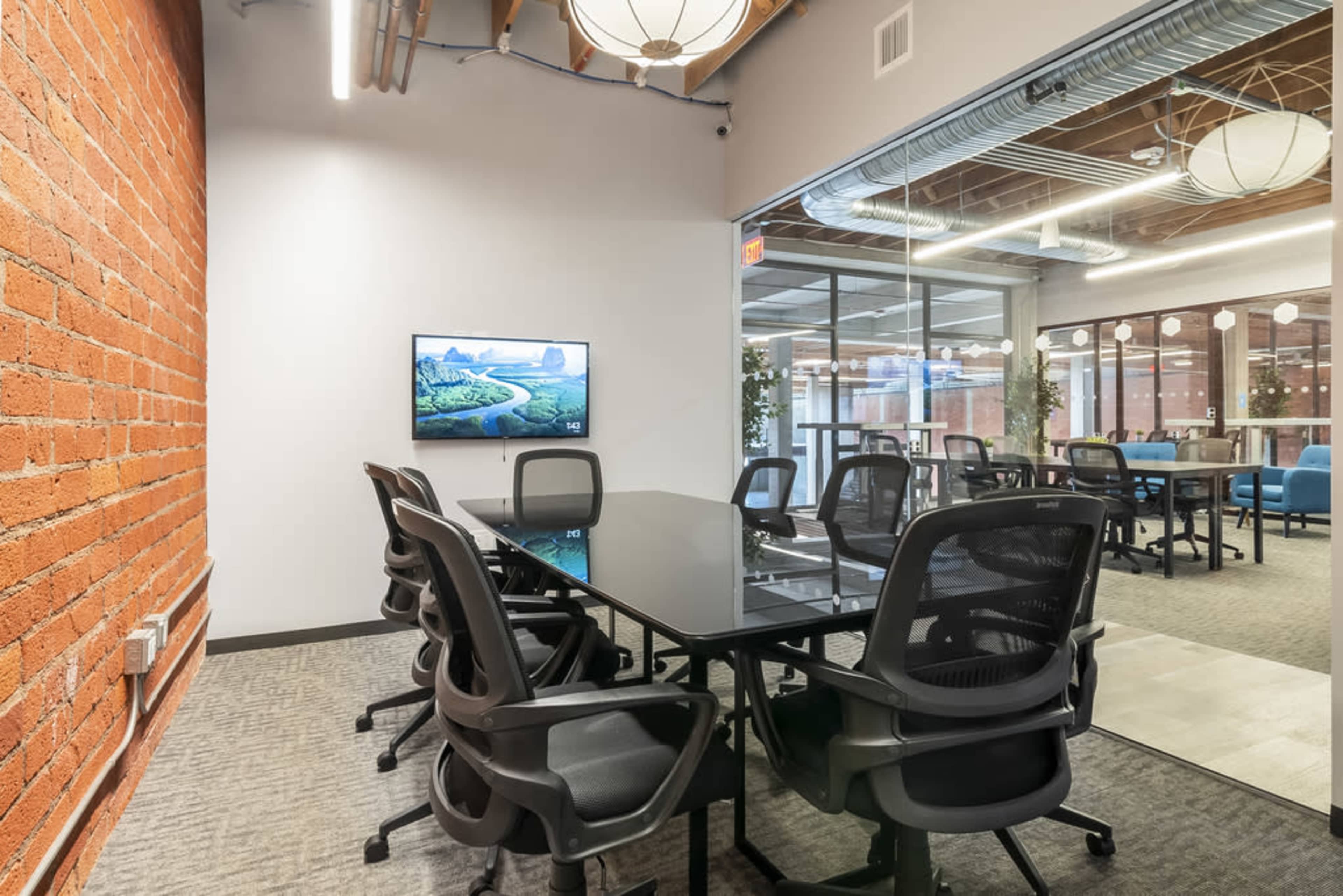 A conference room features a glass table surrounded by black chairs, with a television mounted on a brick wall.