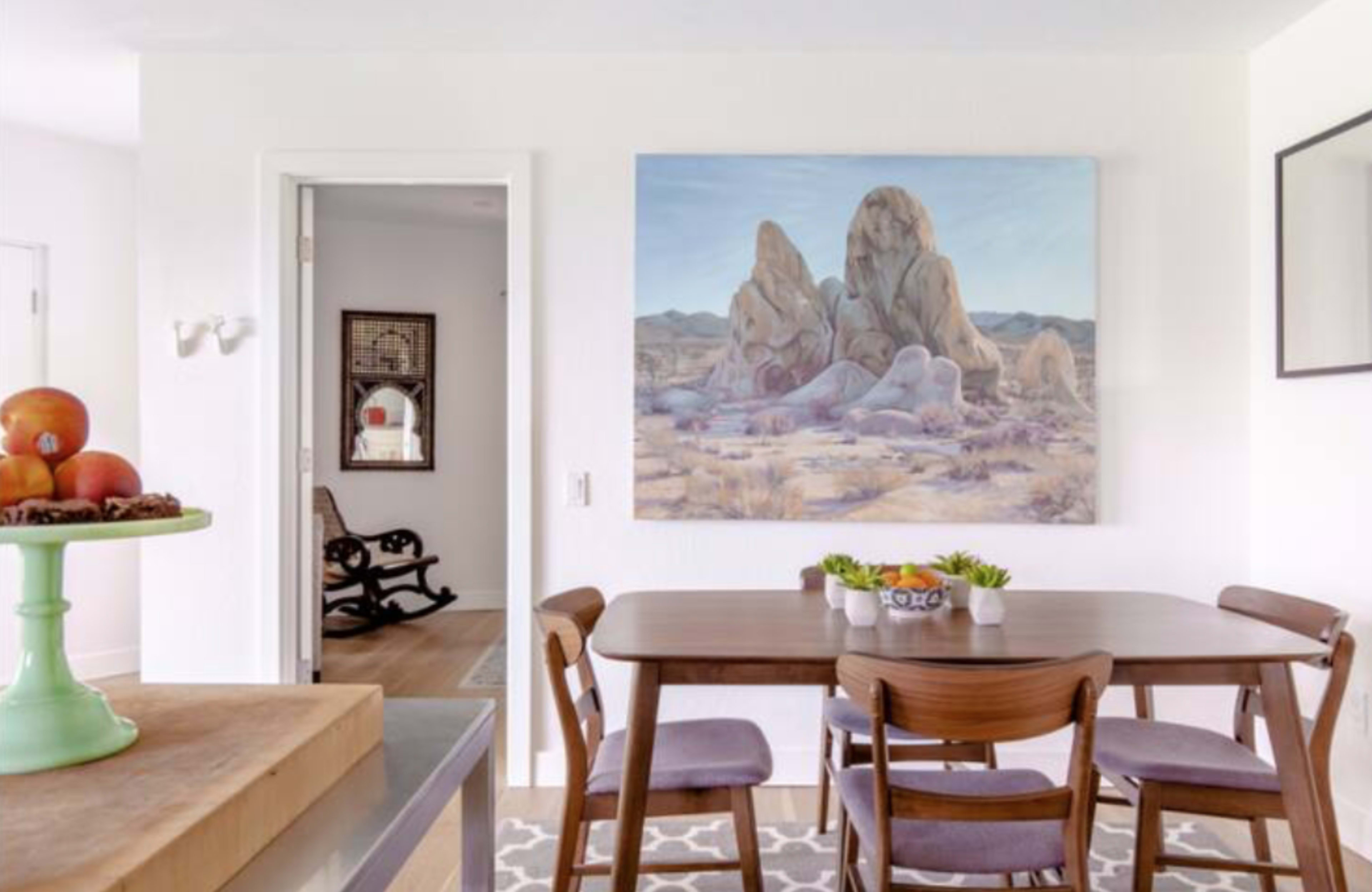 A dining area features a wooden table with four chairs, a fruit bowl on a counter, and a large painting of rocky formations on the wall.