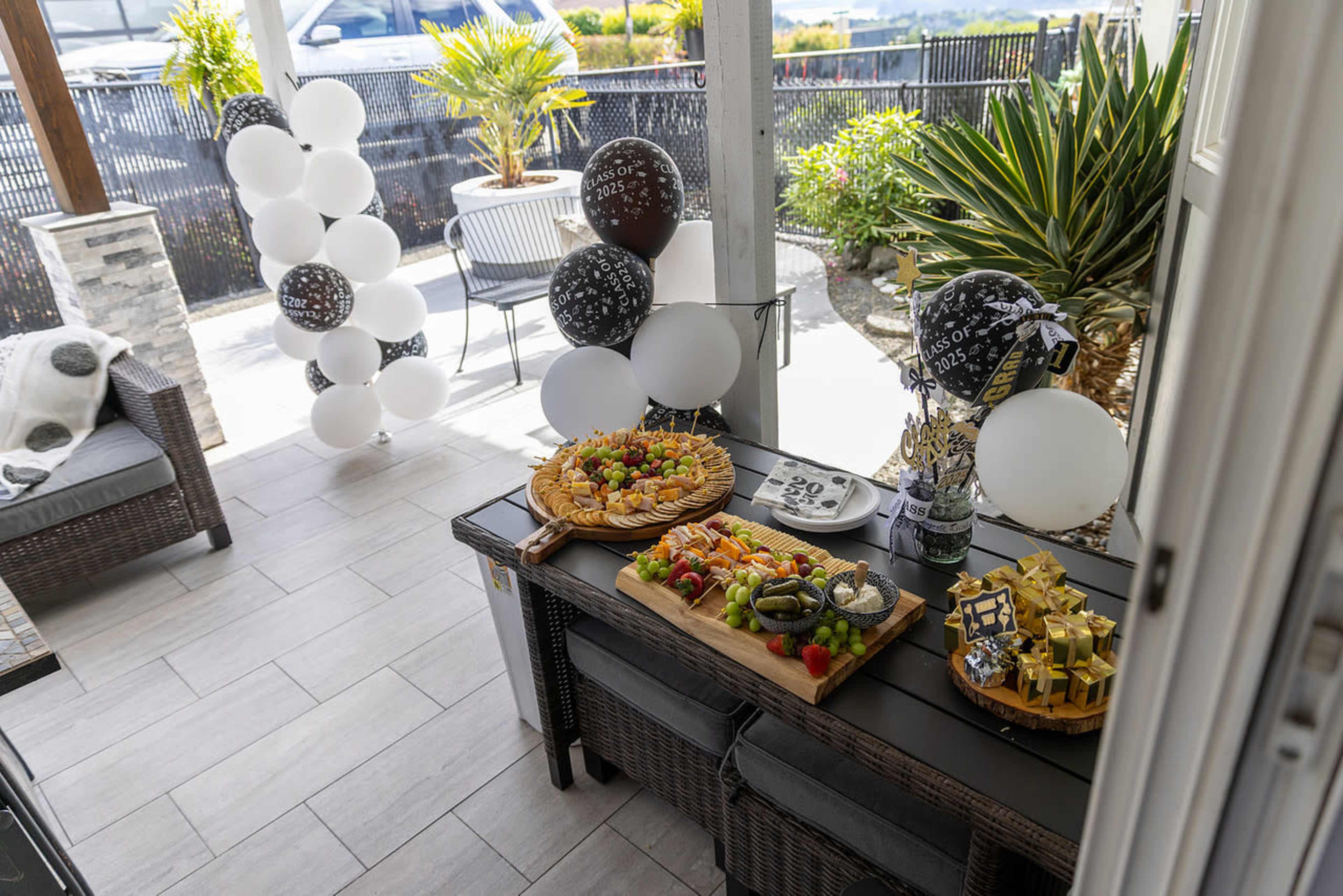 The image shows a decorated outdoor space featuring a table with a variety of colorful food platters, balloons, and plants.