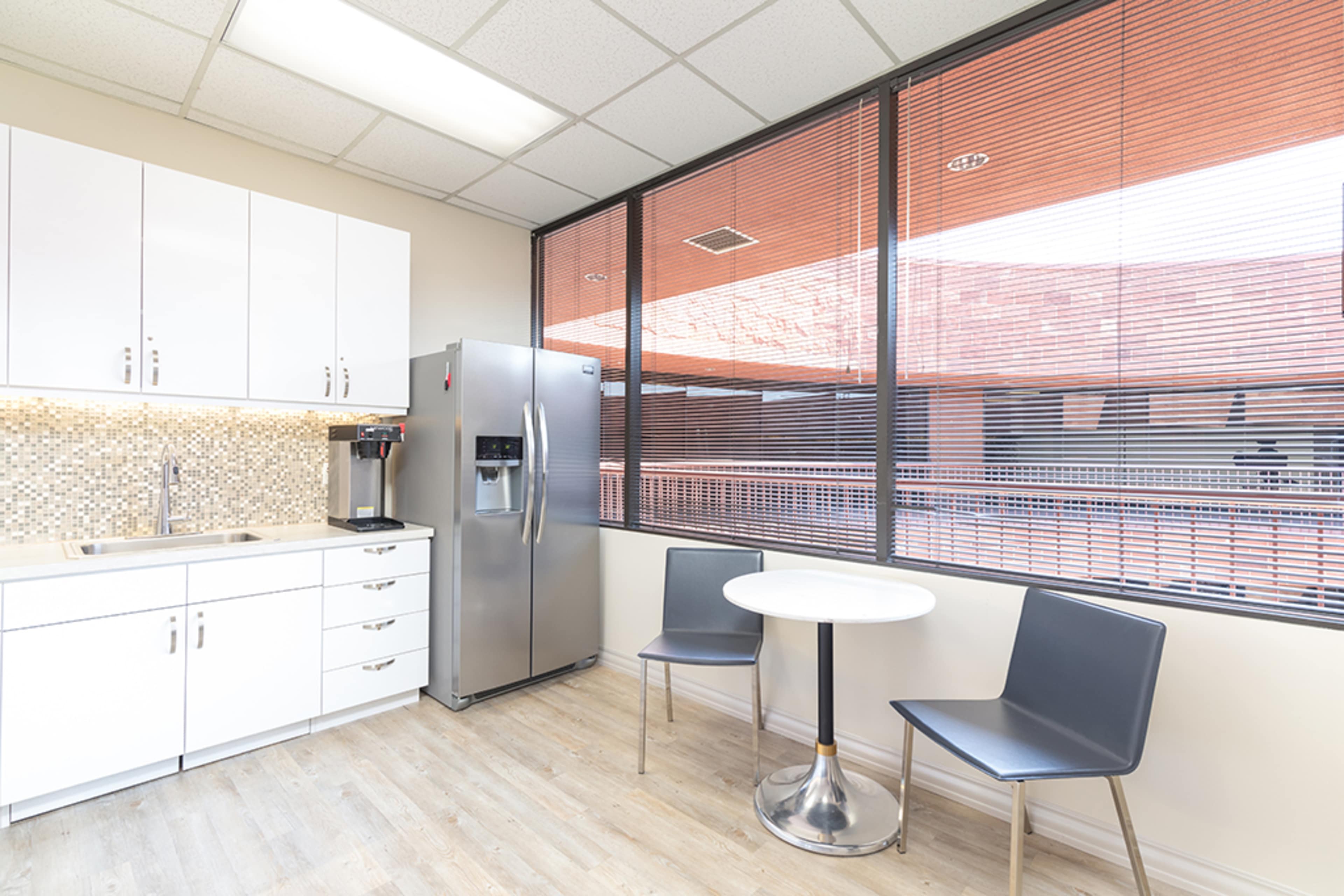 A modern kitchen area features a silver refrigerator, a sink, white cabinetry, and a small round table with two chairs beside large windows.