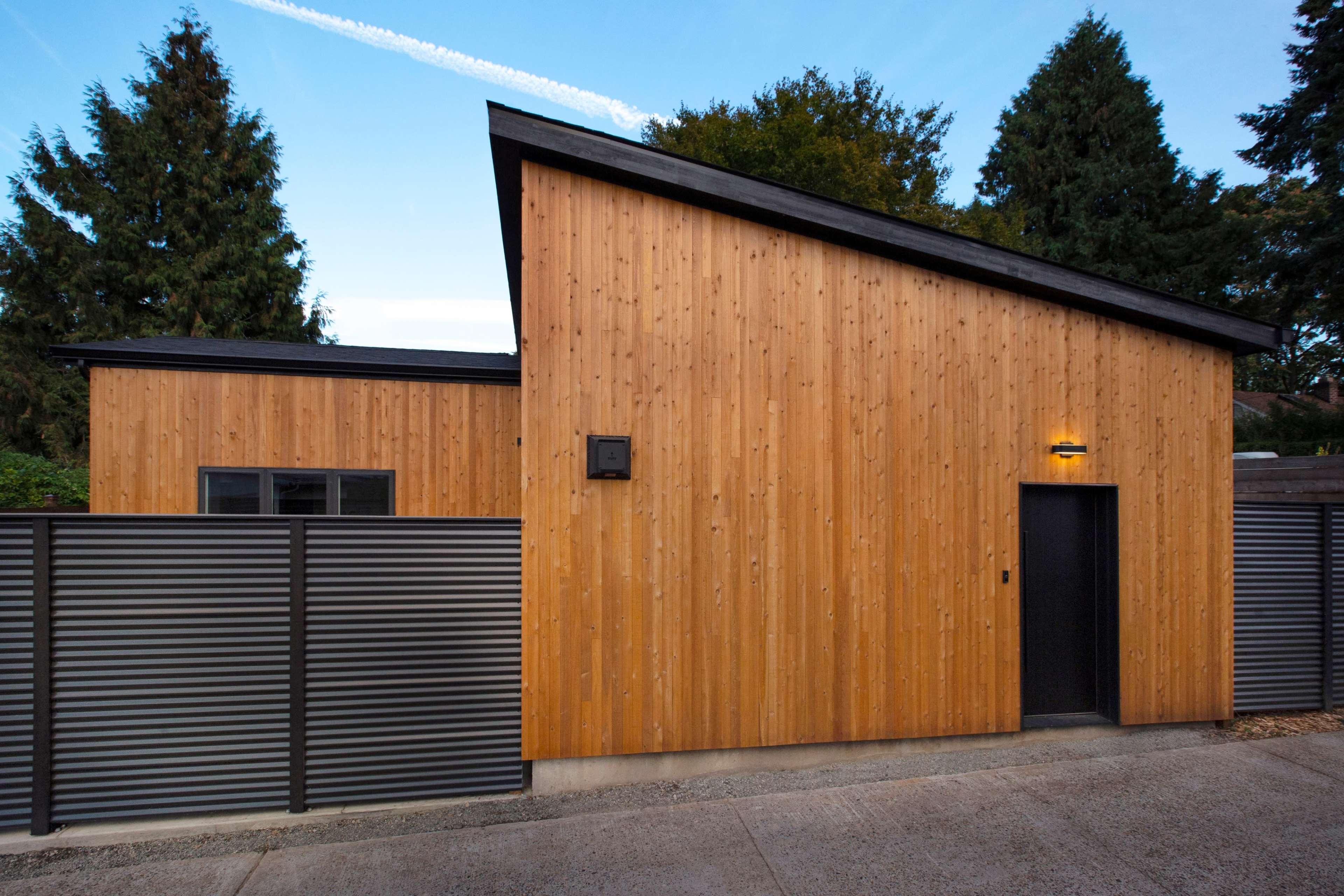 A modern wooden house with a sloped roof and a black door, surrounded by a gray metal fence.