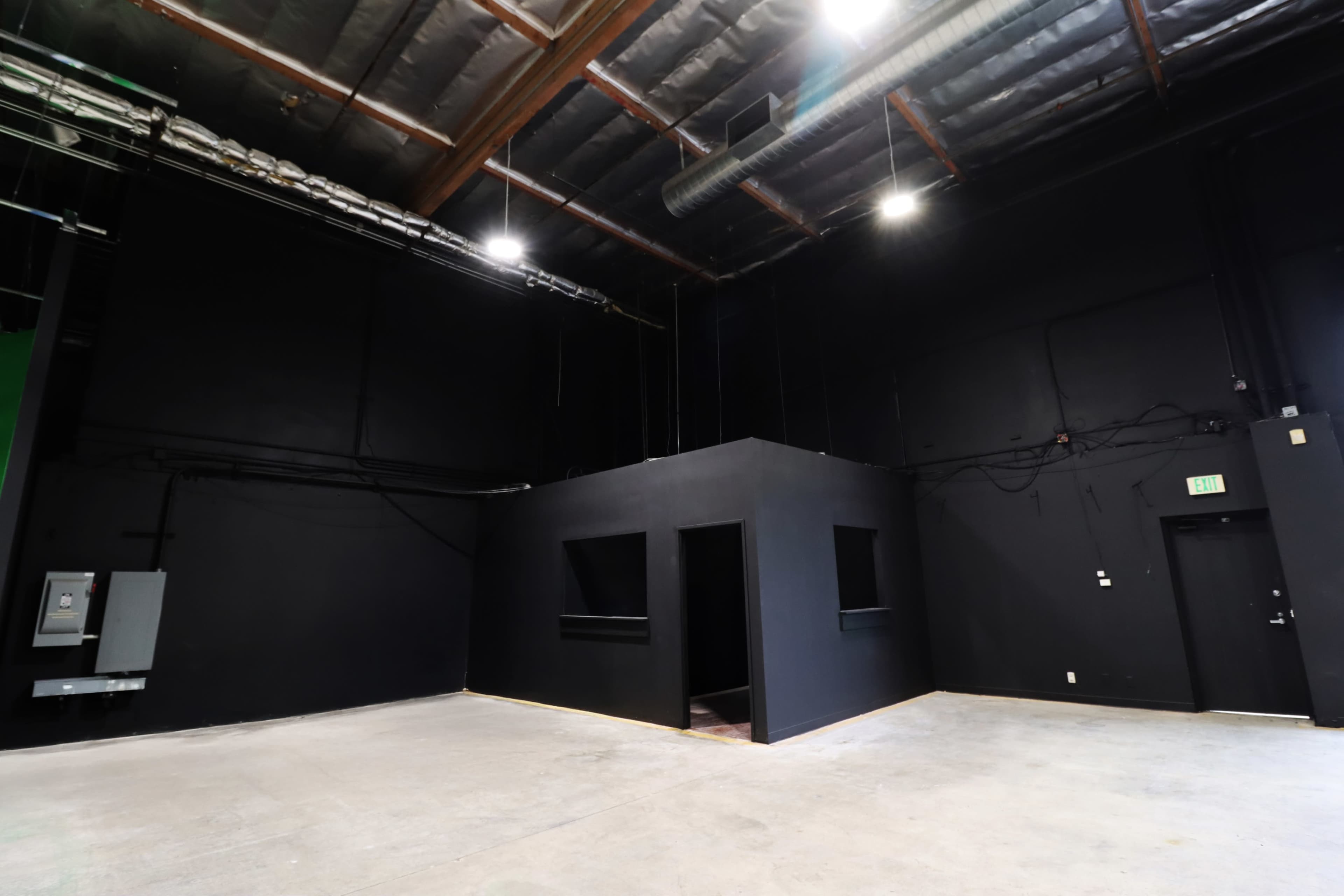 The image shows a spacious, empty room with black walls and a small black structure in the center, featuring two windows and a doorway.