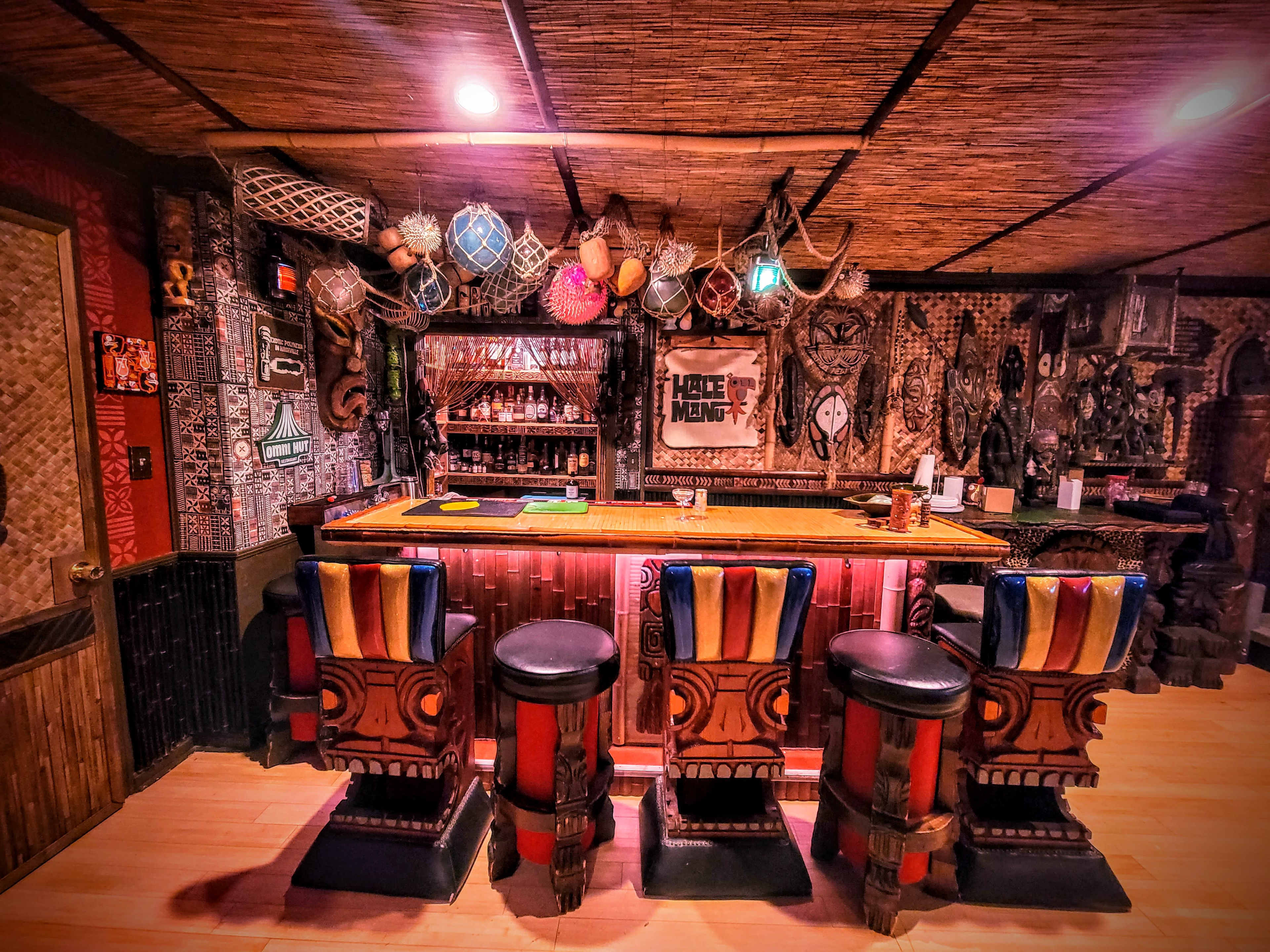The image depicts a tiki-style bar equipped with colorful stools and various decorative artifacts lining the walls and ceiling.