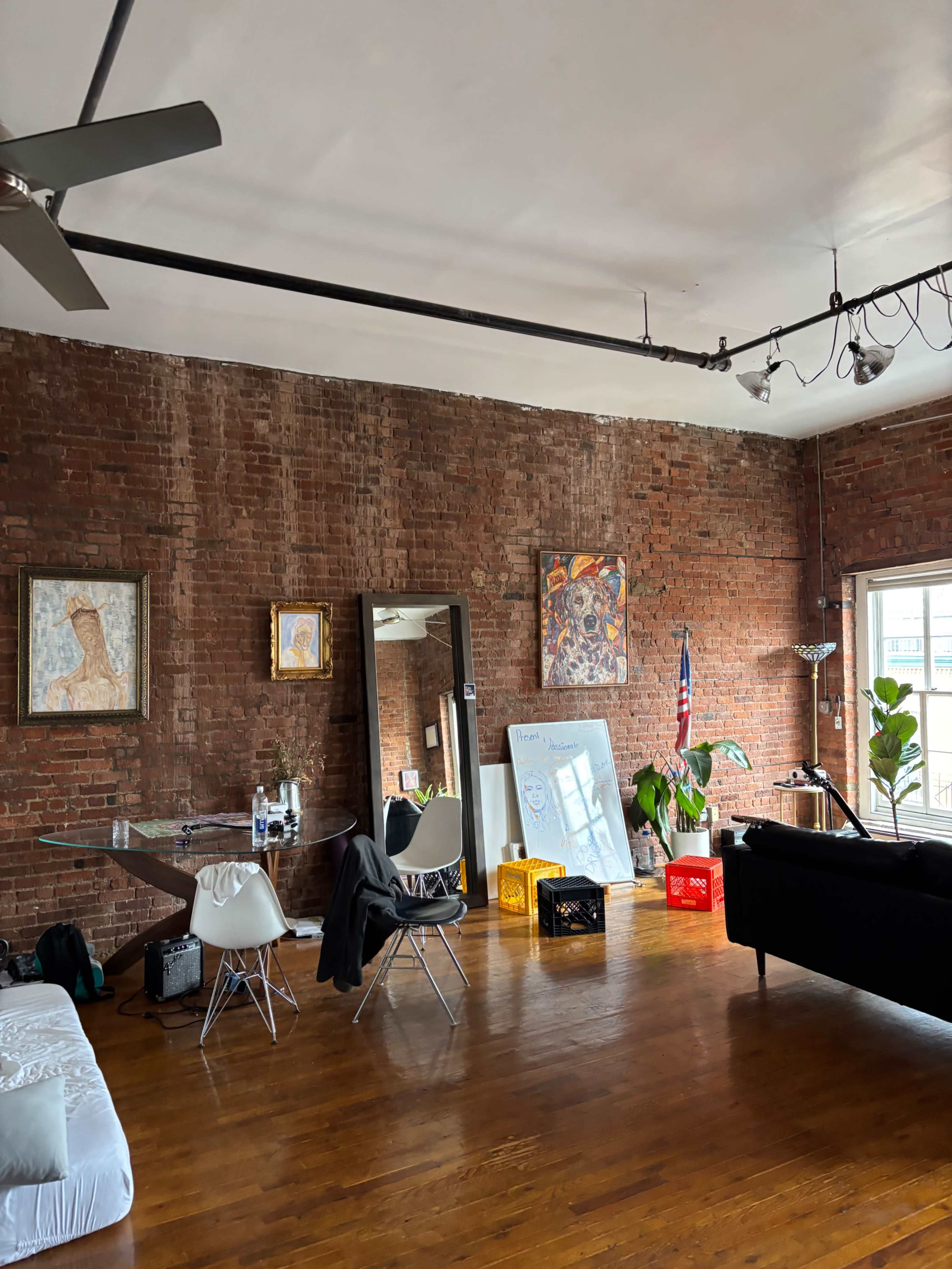 The image shows a spacious room with exposed brick walls, wooden flooring, a dining table with chairs, a large mirror, various artworks, and a sofa by a window.