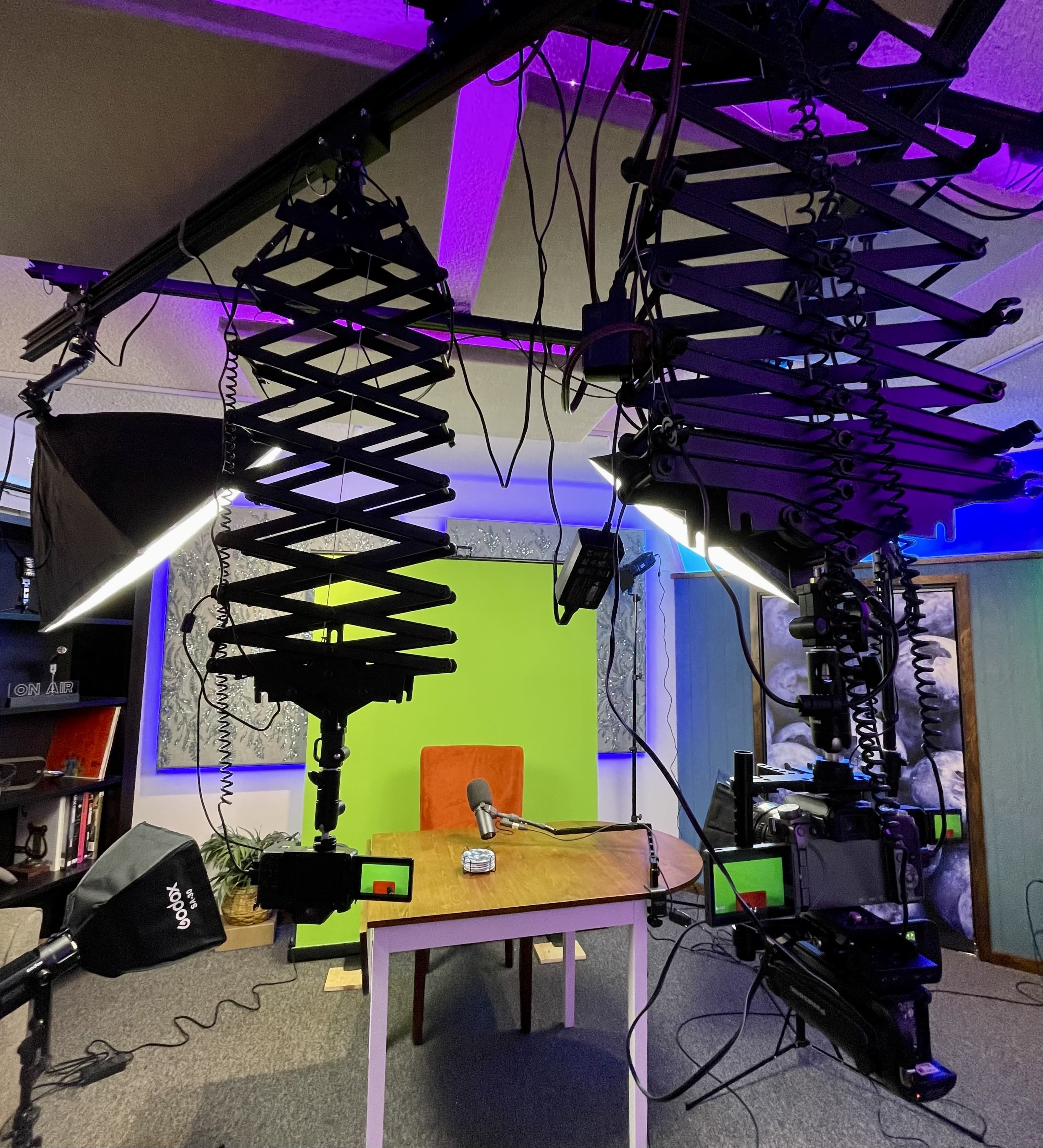 The image shows a video recording setup with two tall light stands, a table in the center, and a bright green backdrop, all surrounded by various camera equipment and lighting.