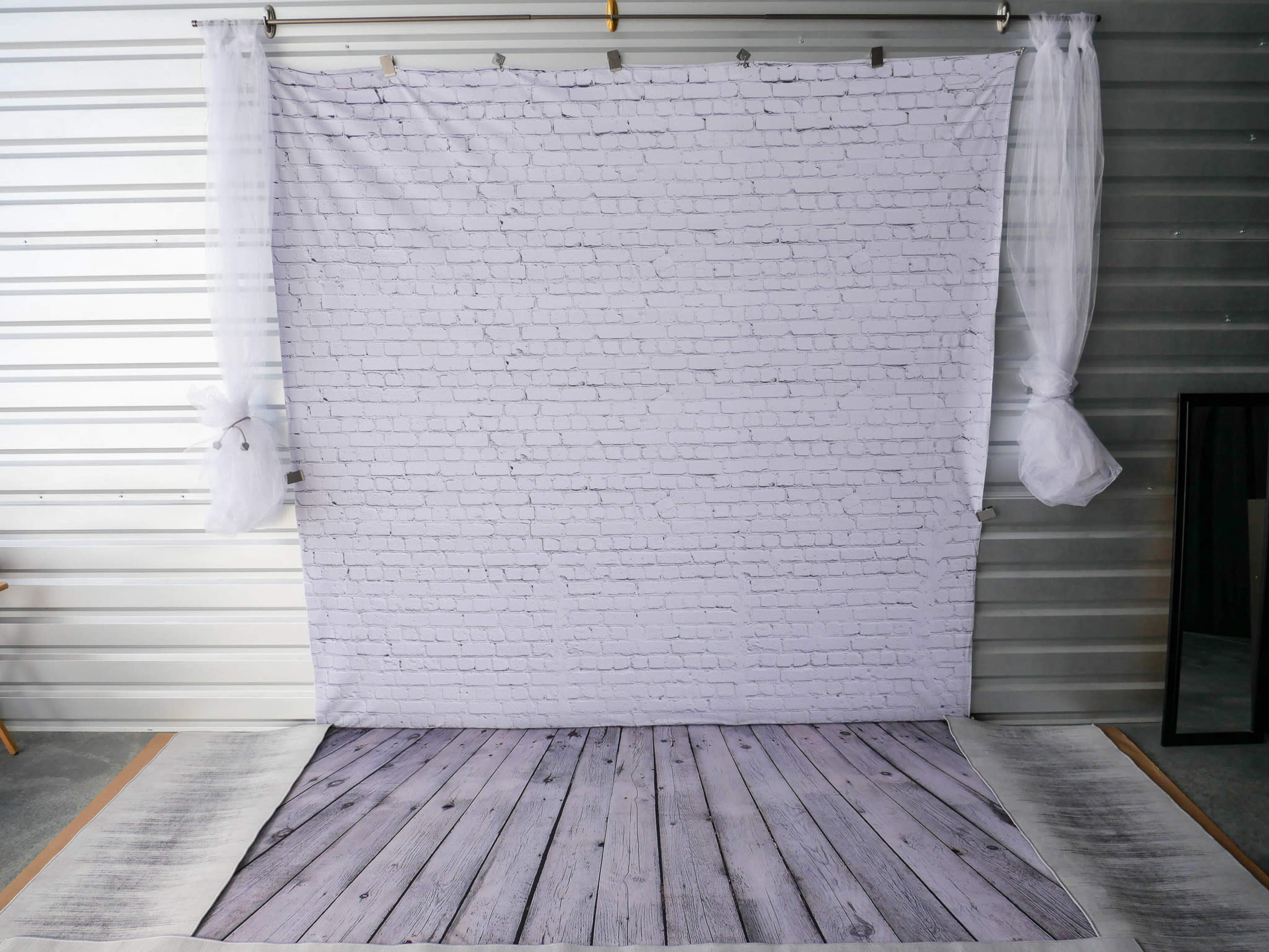 A backdrop featuring a white brick pattern alongside a wooden floor pattern, set in a studio space with sheer curtains and a mirror.