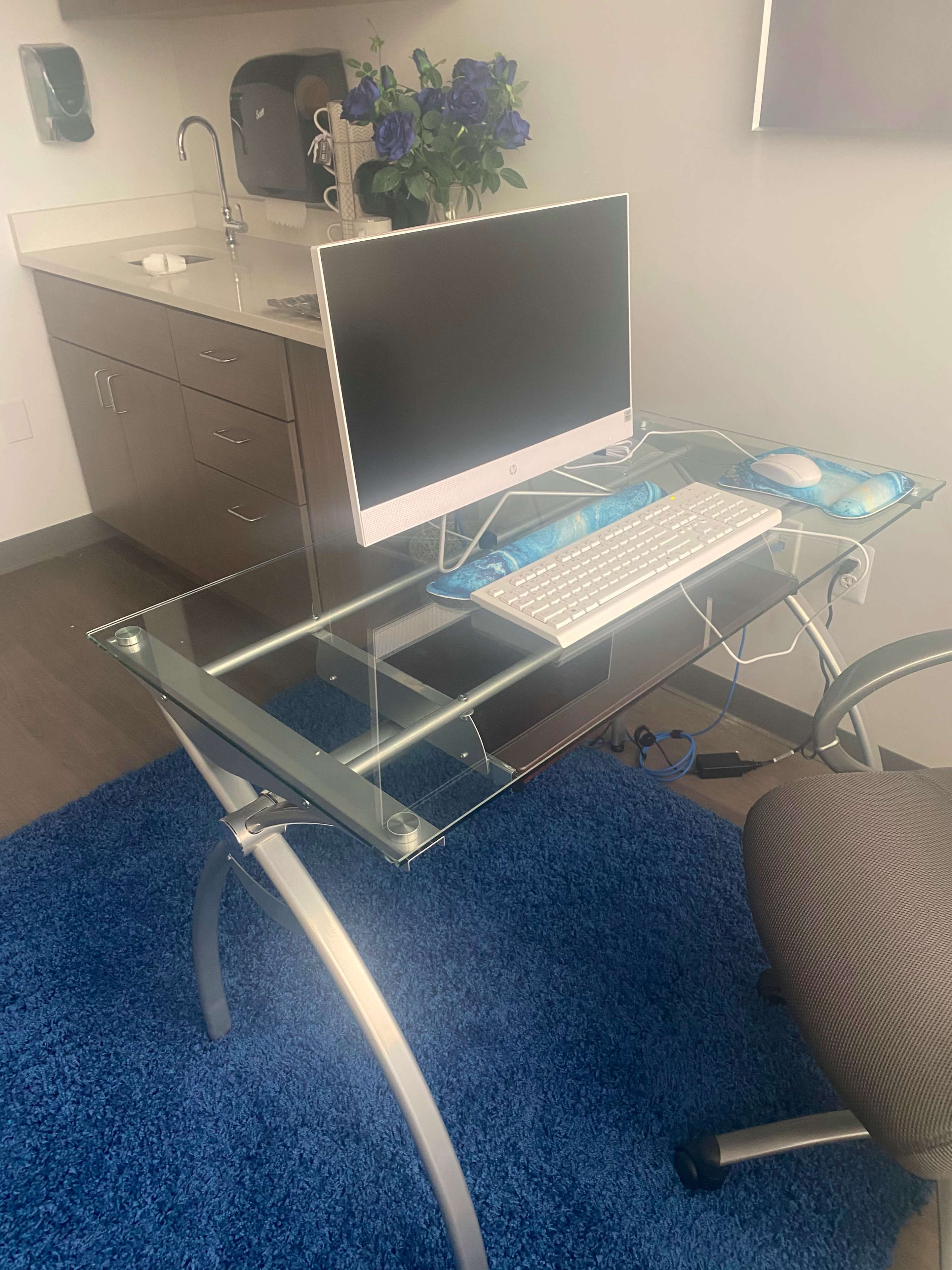 A glass computer desk with a monitor, keyboard, and mouse is positioned in front of a small kitchen area.