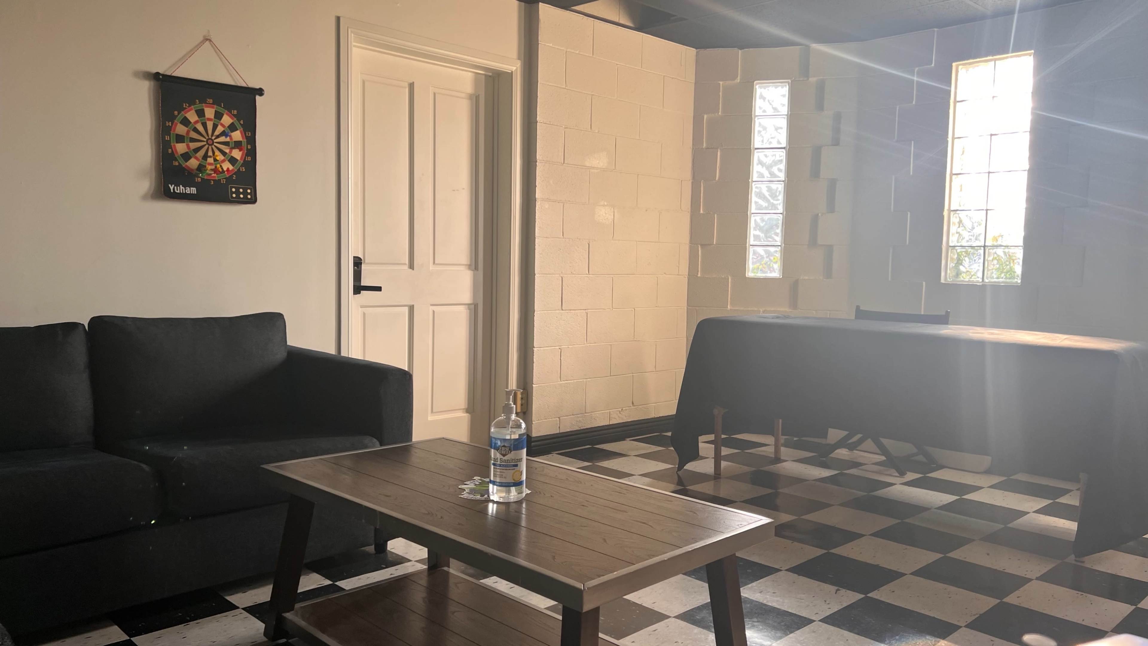 A dimly lit room features a black couch, a wooden table with a bottle on it, and a dartboard hanging on the wall.
