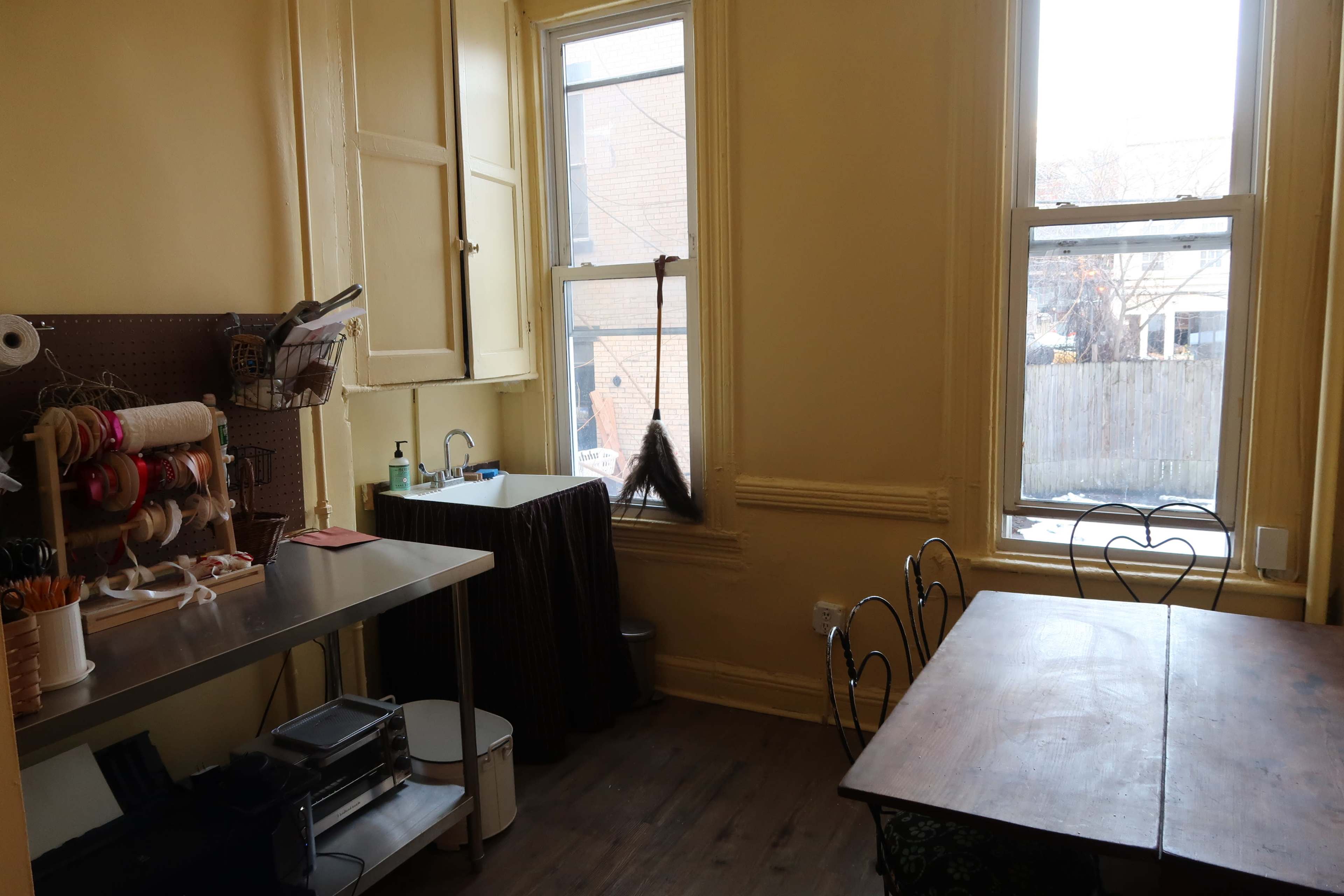 The image depicts a small, bright room with a metal table, a sink, a window with a broom leaning against it, and a wooden dining table with metal chairs.