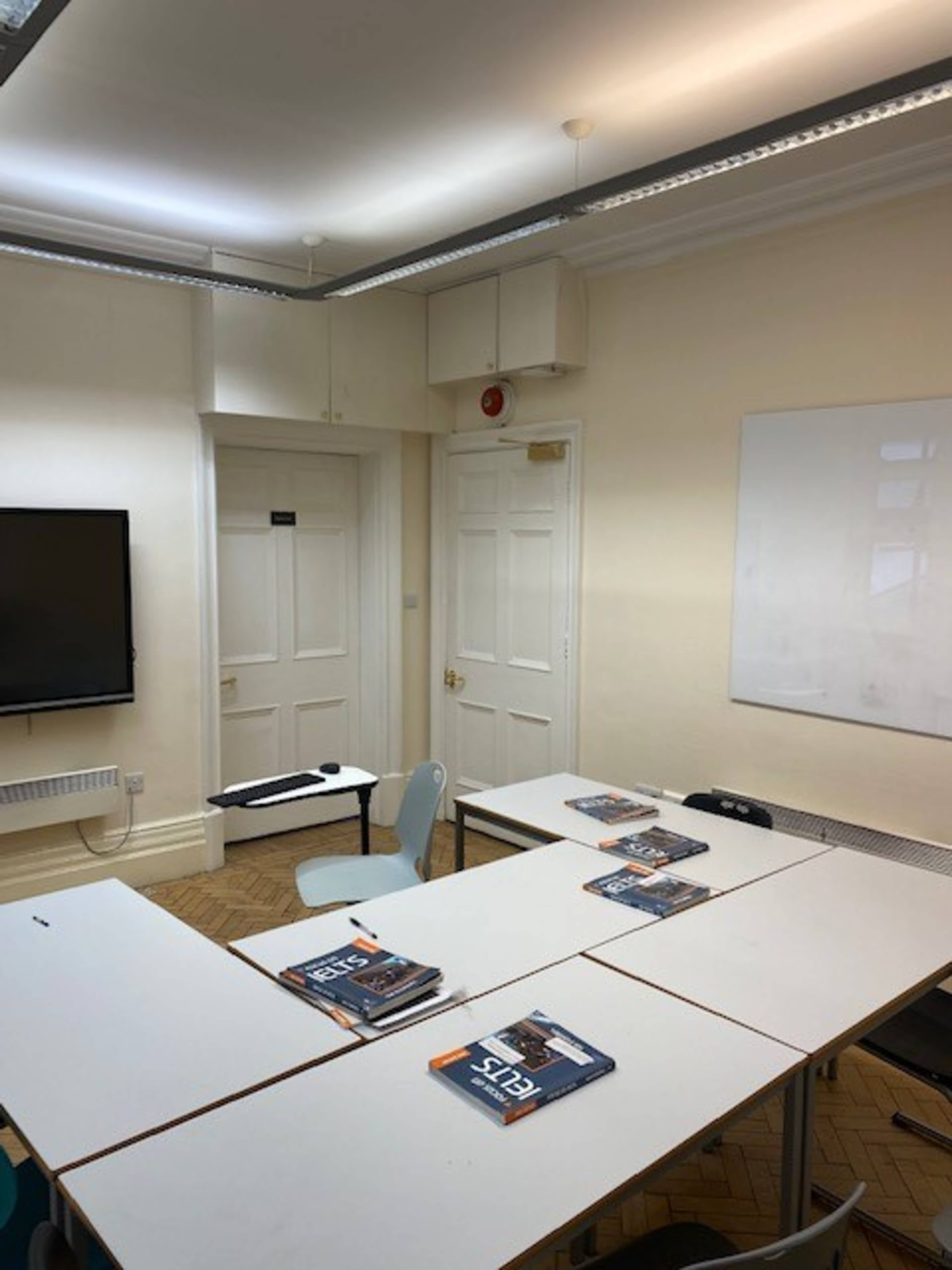 A classroom with several tables arranged in a U-shape, each holding textbooks, along with a television and a whiteboard on the walls.