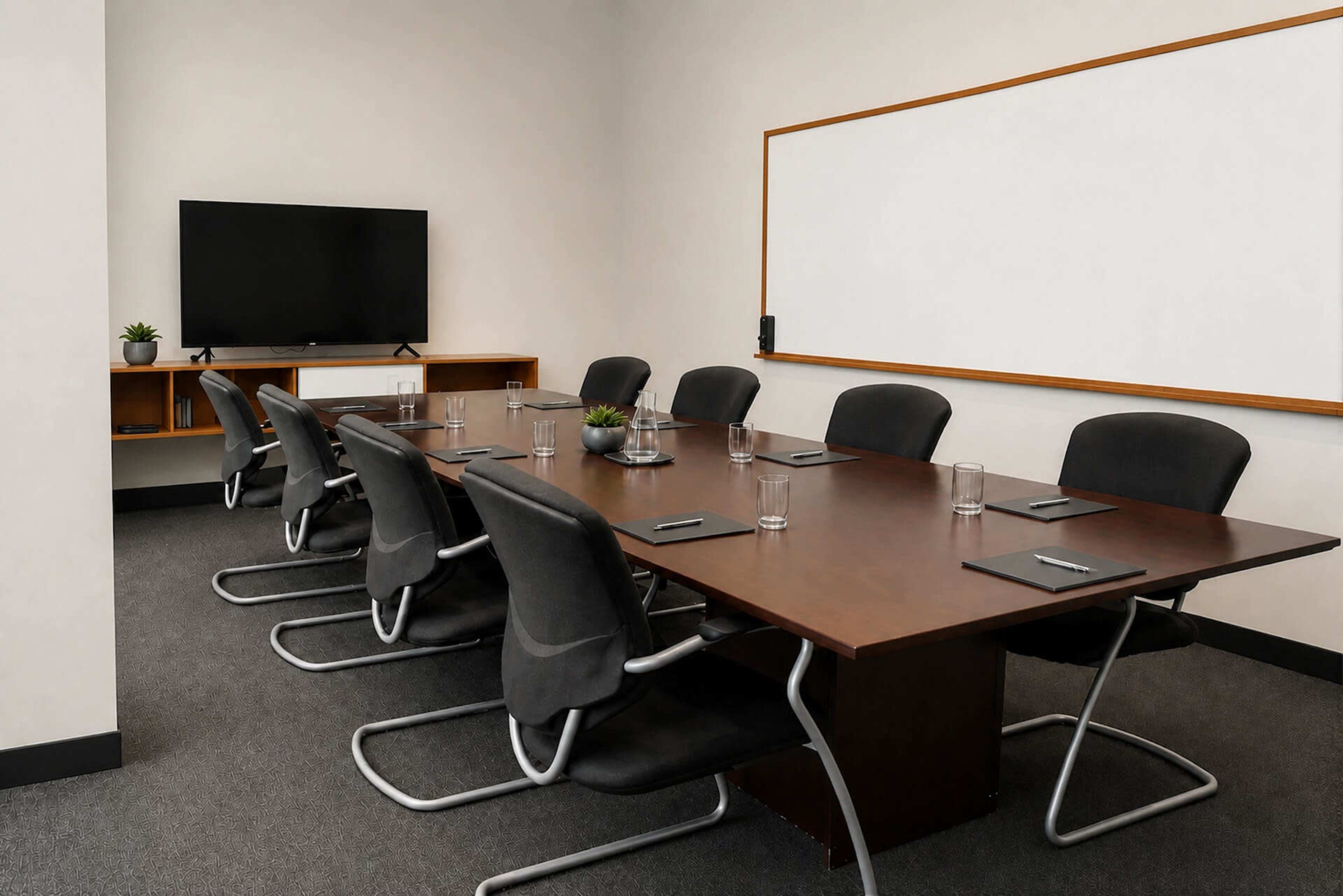 A conference room features a large wooden table surrounded by ergonomic chairs, with a television mounted on one wall and a whiteboard on another.