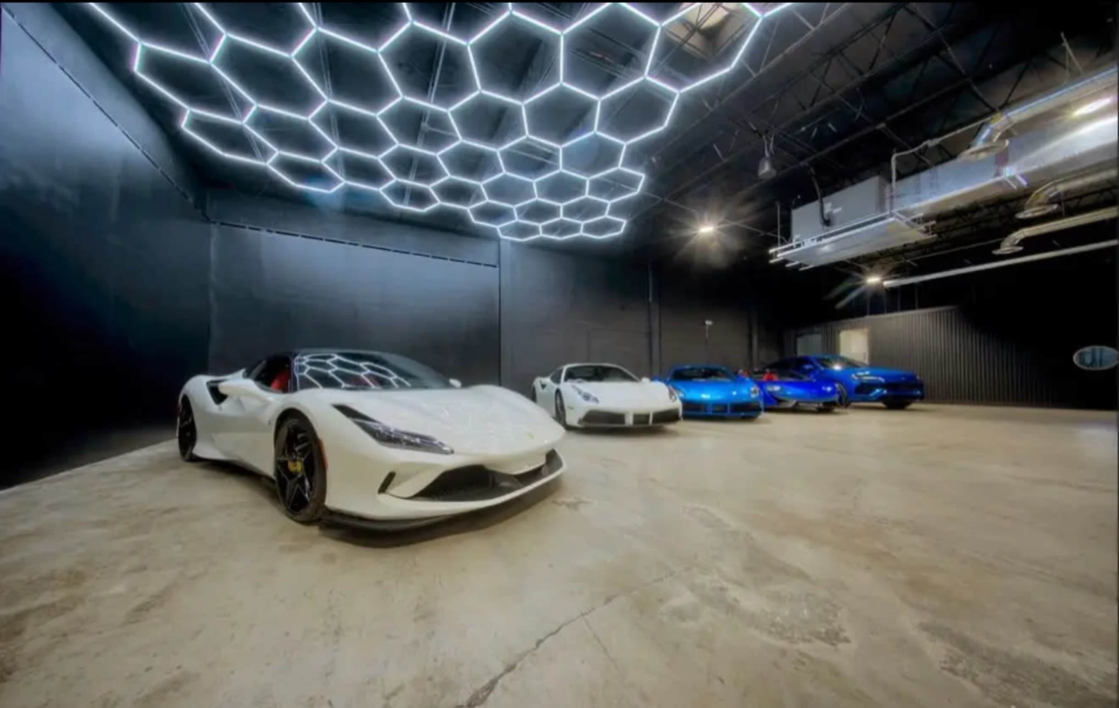 A row of luxury sports cars, including white and blue models, is displayed in a sleek, modern showroom with a geometric light installation on the ceiling.