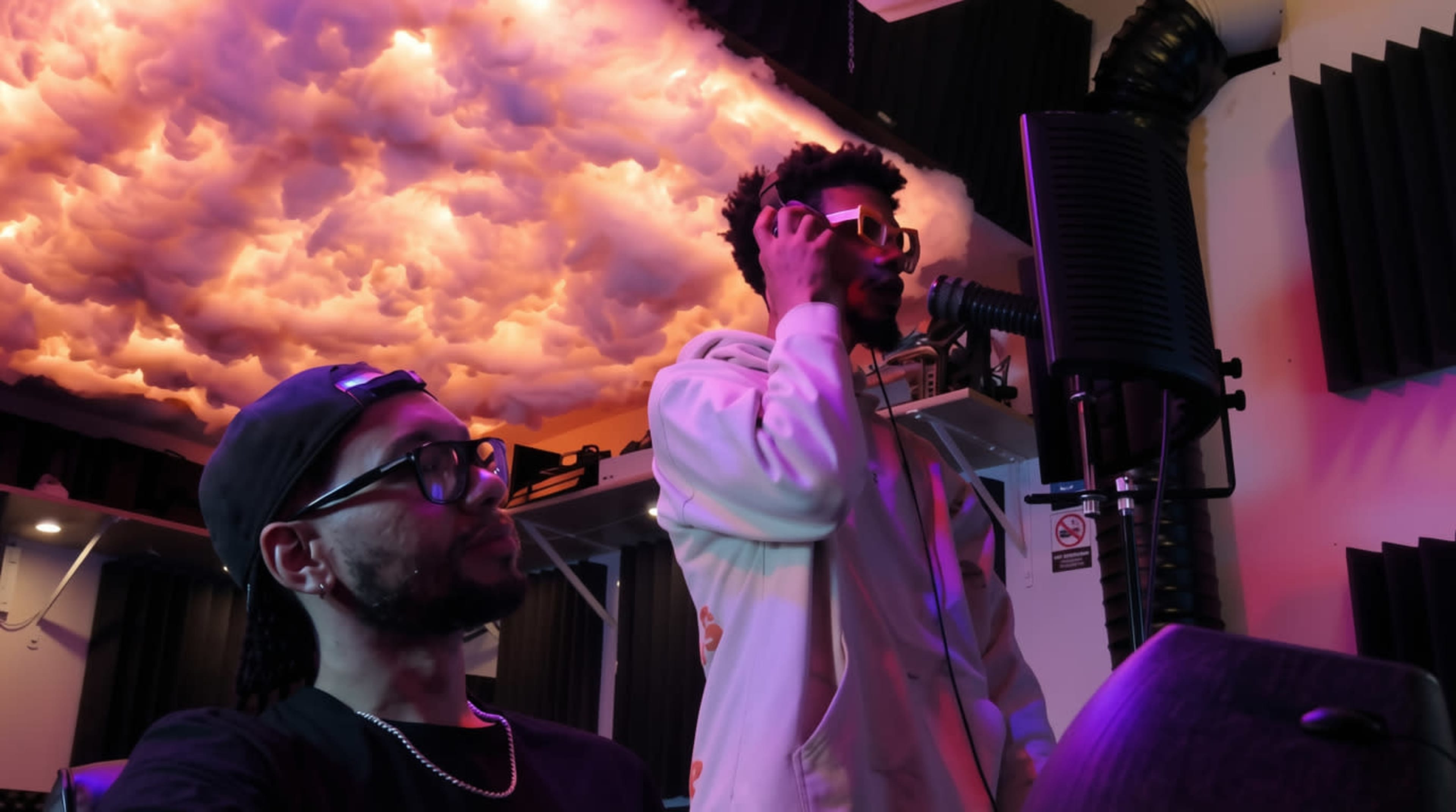 Two individuals are present in a recording studio, with one person standing and singing into a microphone while the other sits nearby, observing.