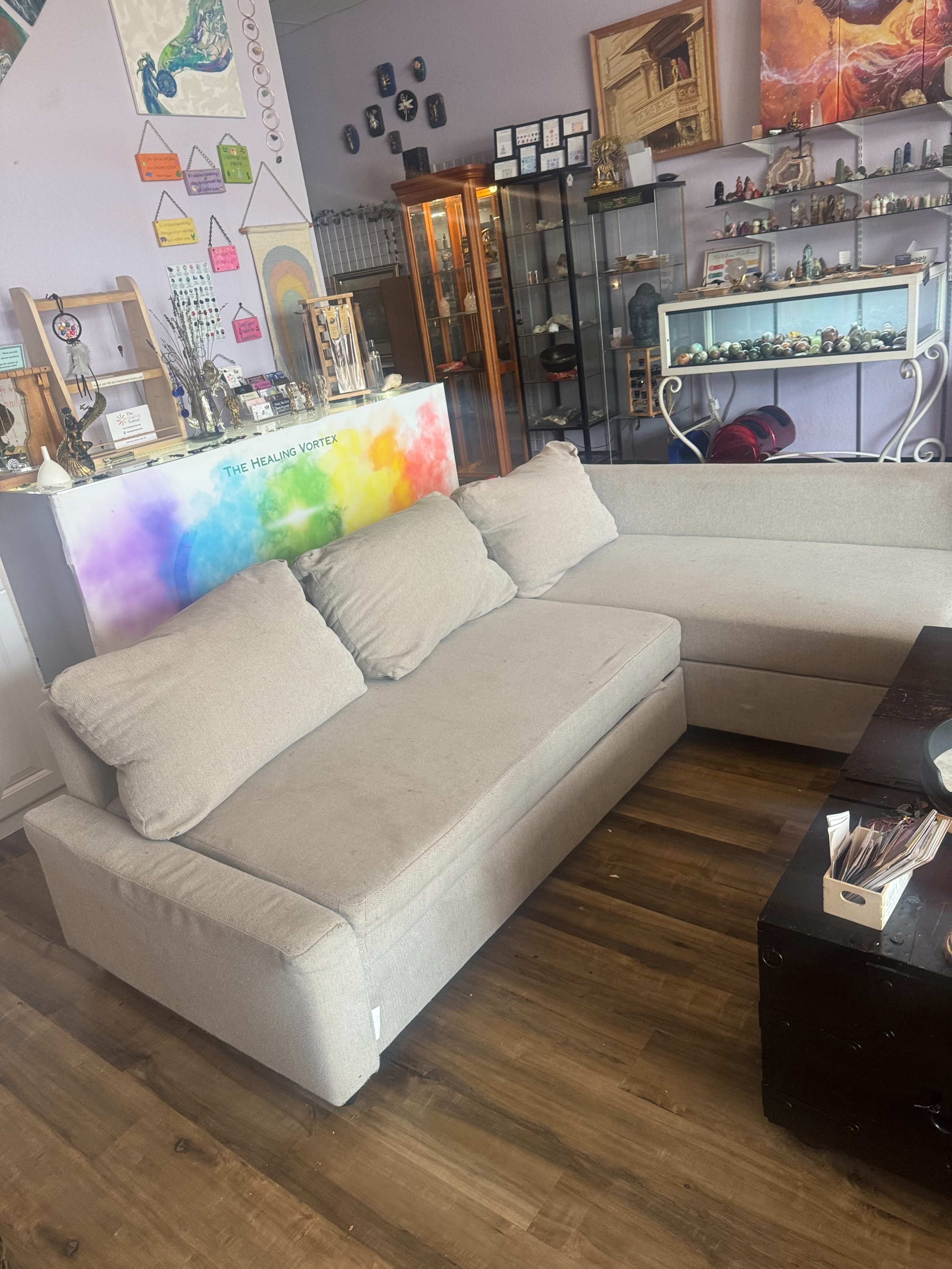 The image shows a light-colored sectional sofa with two large cushions, placed in a room featuring shelves with various decorative items and a colorful display behind the sofa.