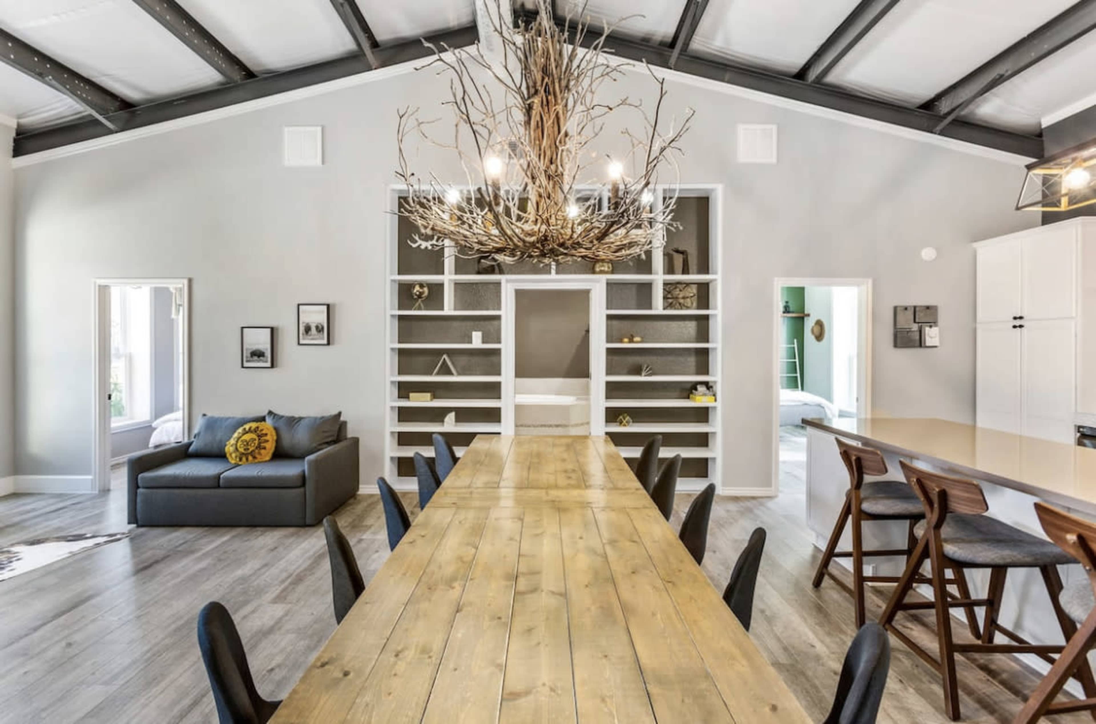 A spacious dining area featuring a long wooden table surrounded by black chairs, with a large chandelier made of branches overhead and a cozy seating area to the side.