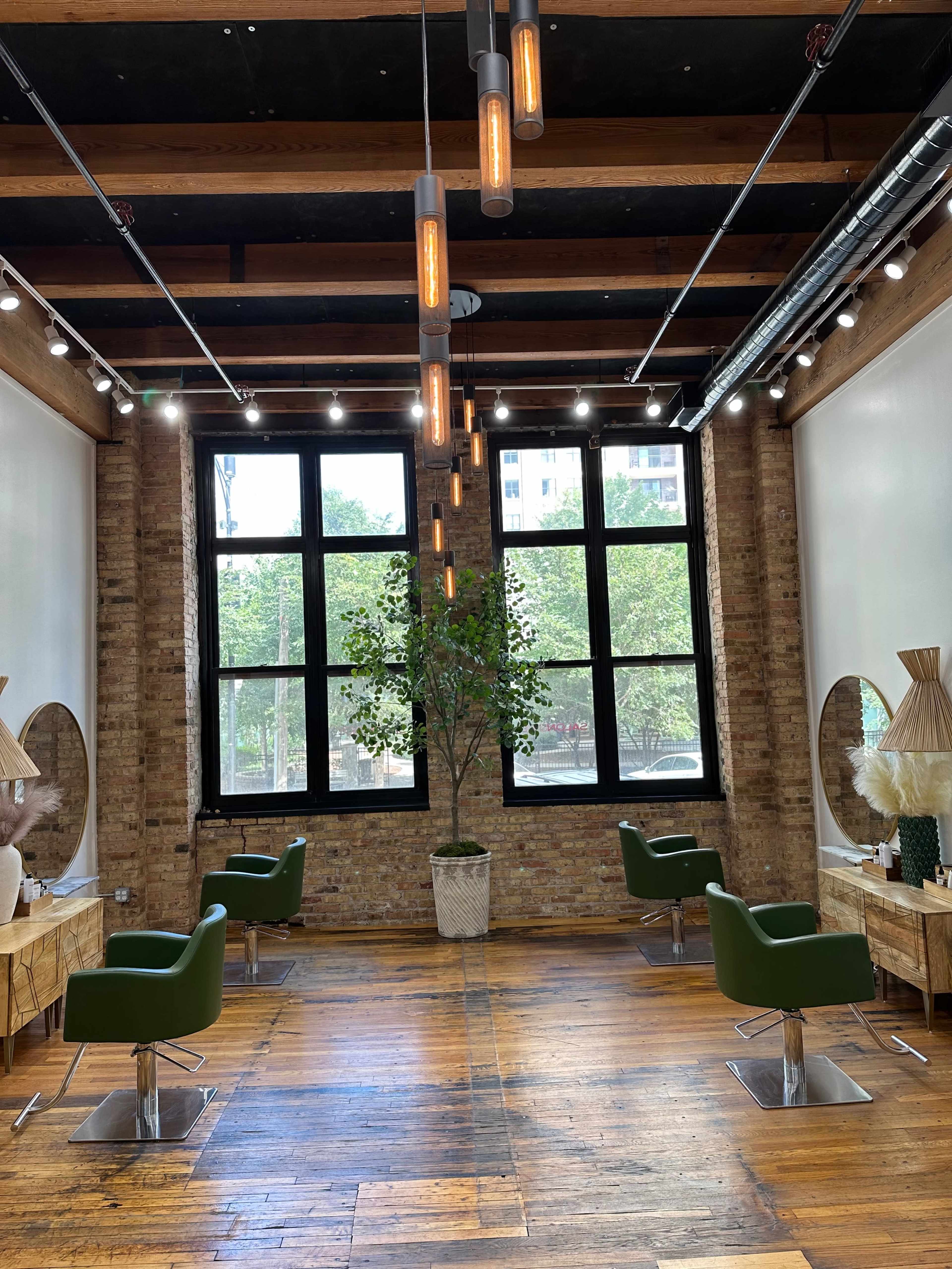 A salon interior featuring two green chairs, large windows with natural light, and decorative elements including plants and mirrors on wooden floors.